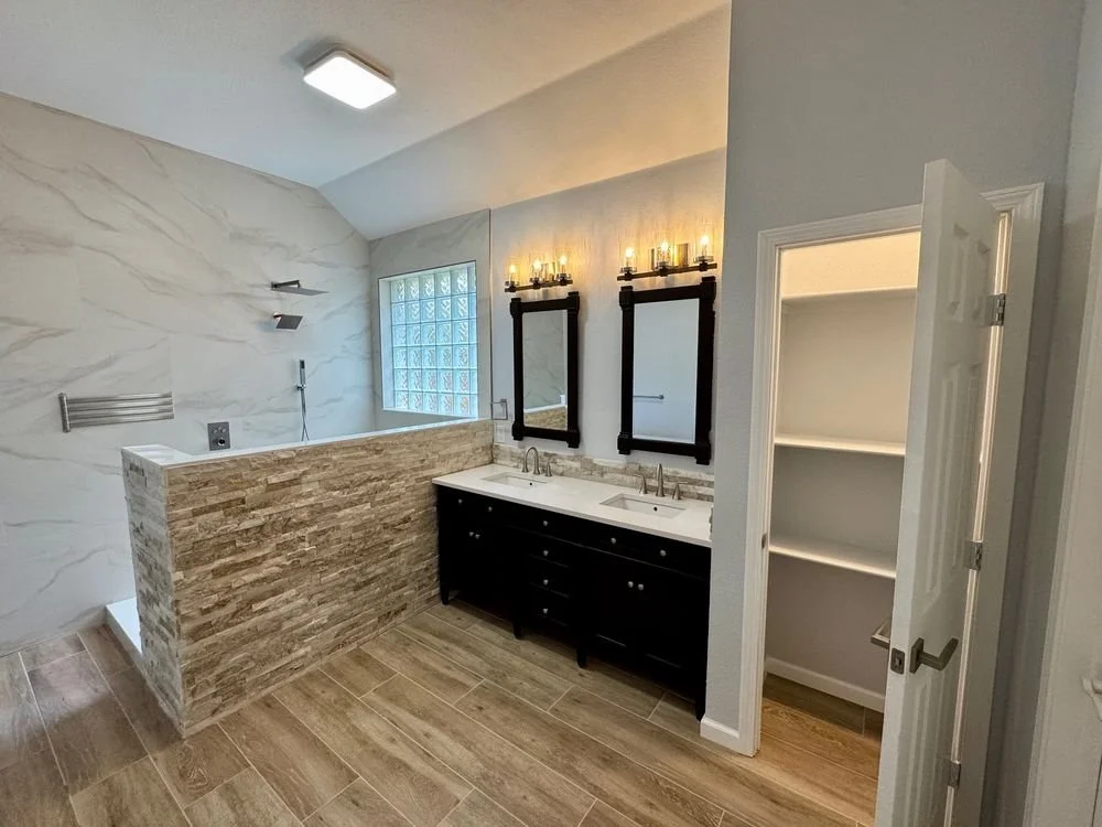Bathroom with double sink vanity, black-framed mirrors, textured wall above the sinks, shelves, a glass block window, and a walk-in shower with stone tiles on the half wall.