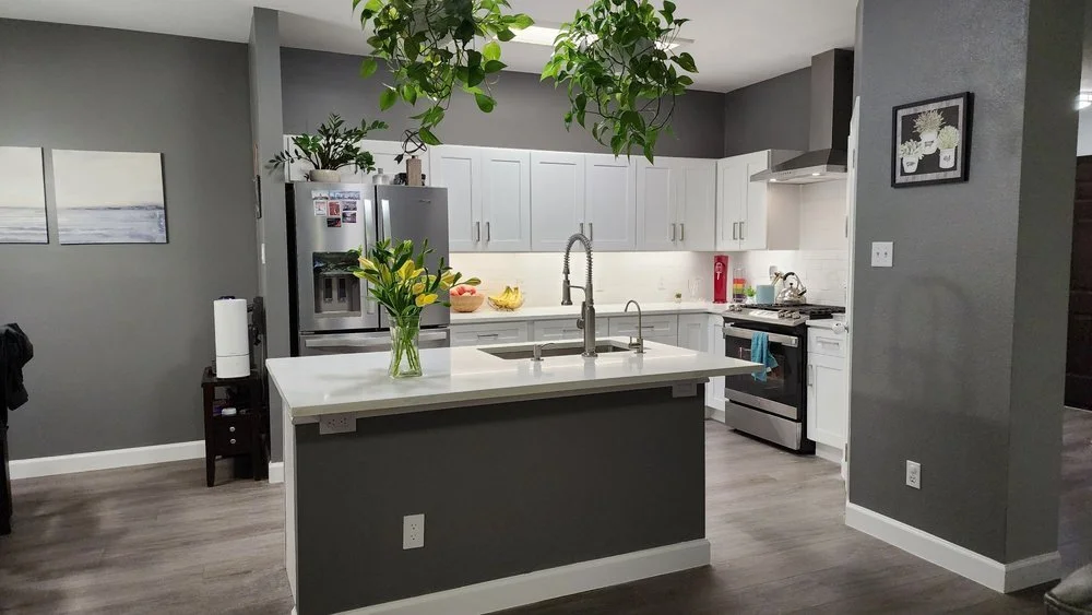 Modern kitchen with white cabinets, gray walls, and a central island with a vase of flowers. There is stainless steel refrigerator, stove, and a sink with a high-arc faucet. Green plants hang from the ceiling and are on top of the refrigerator, with 