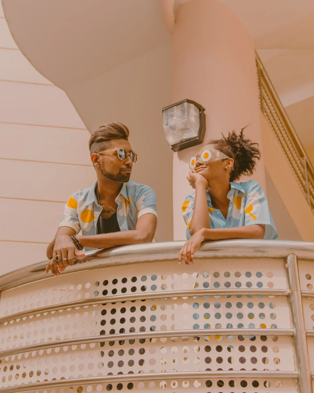 A man and a woman leaning on a railing indoors, both wearing matching blue and yellow polka dot shirts. The woman is wearing eye-shaped glasses with fried egg details, and the man is wearing sunglasses, both looking at each other.