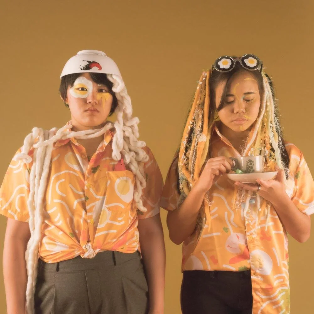 Two women wearing matching patterned shirts with creative makeup and accessories, one with a bowl and the other with an upside-down bowl on her head, posing against a yellow background.