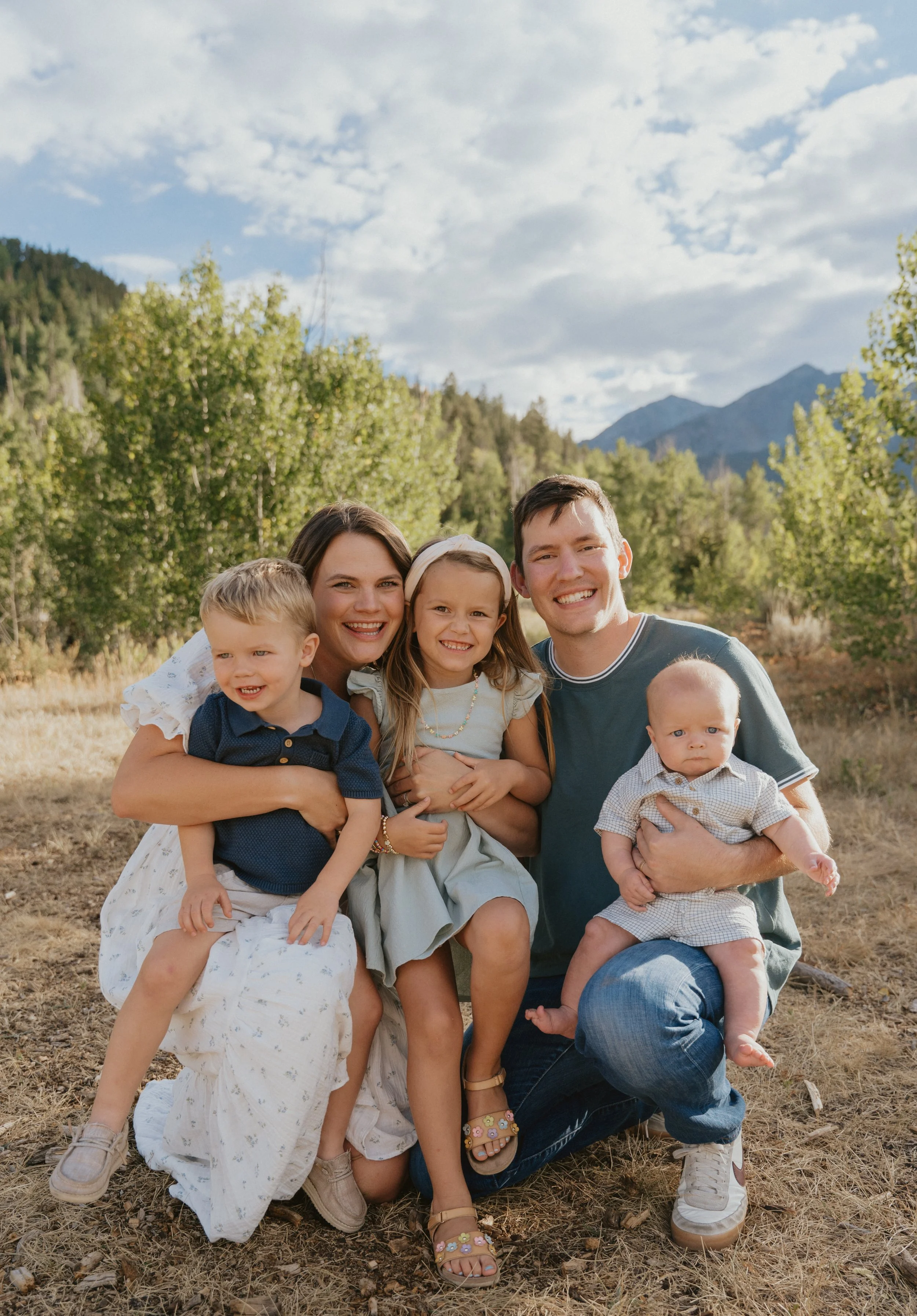 A happy family of six, including two adults and four children, outdoors in a scenic natural setting with mountains and trees in the background.