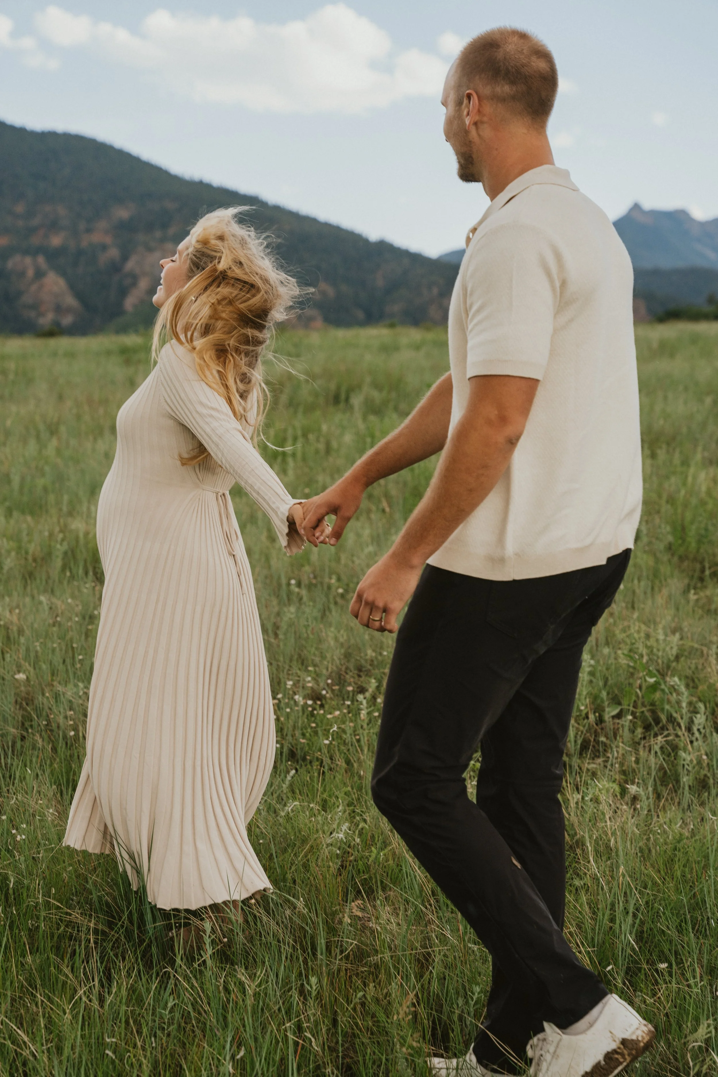 A couple holding hands and dancing or walking in a grassy field, with mountains and a partly cloudy sky in the background.