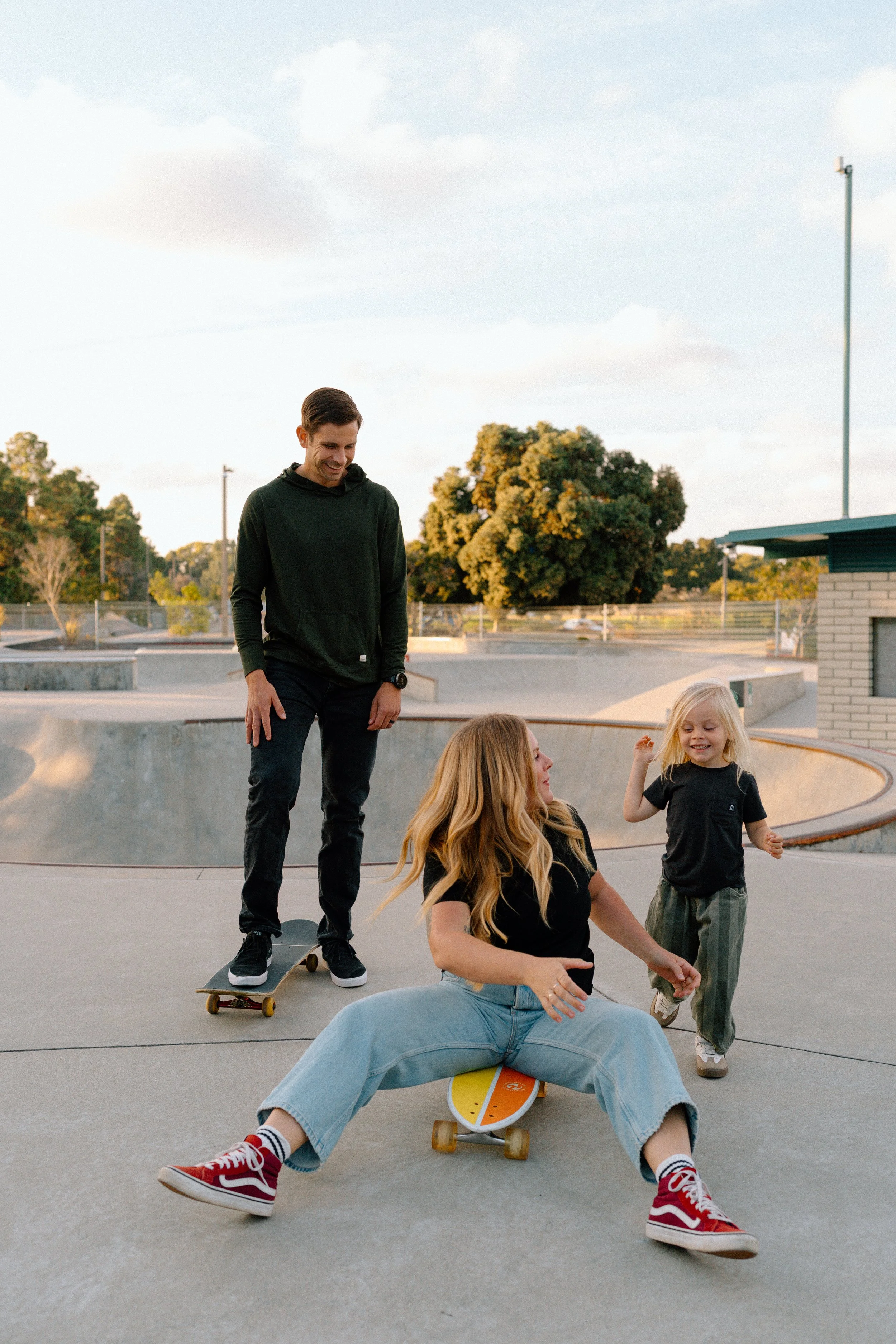 A family at a skatepark enjoying skateboarding together. The mother is sitting on a skateboard, the father is standing on a skateboard, and a young girl is walking nearby, all smiling and having fun.