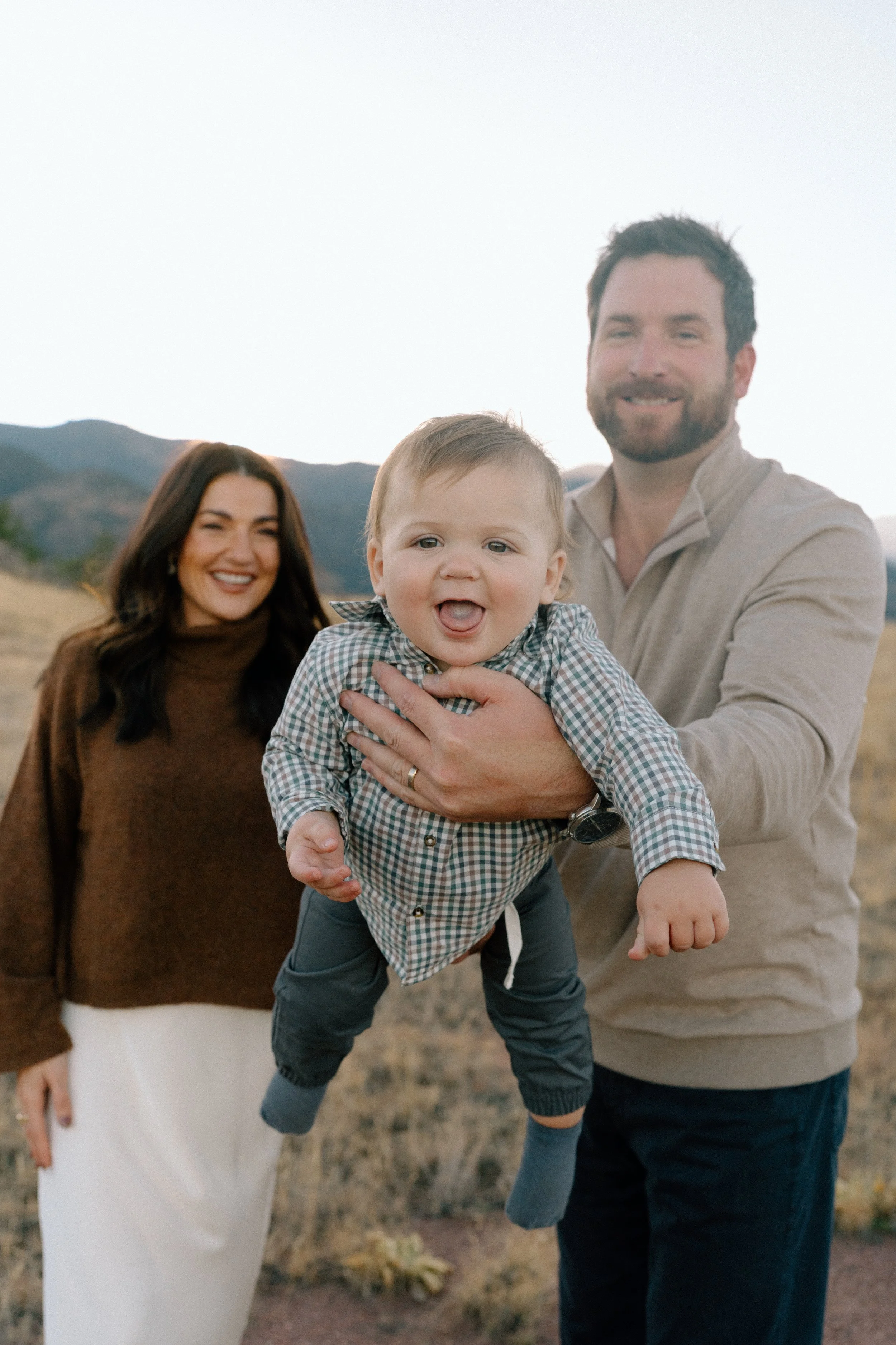 A smiling family of three outdoors with a mountain landscape in the background. A man holds a young boy, and a woman stands nearby, all appearing happy.