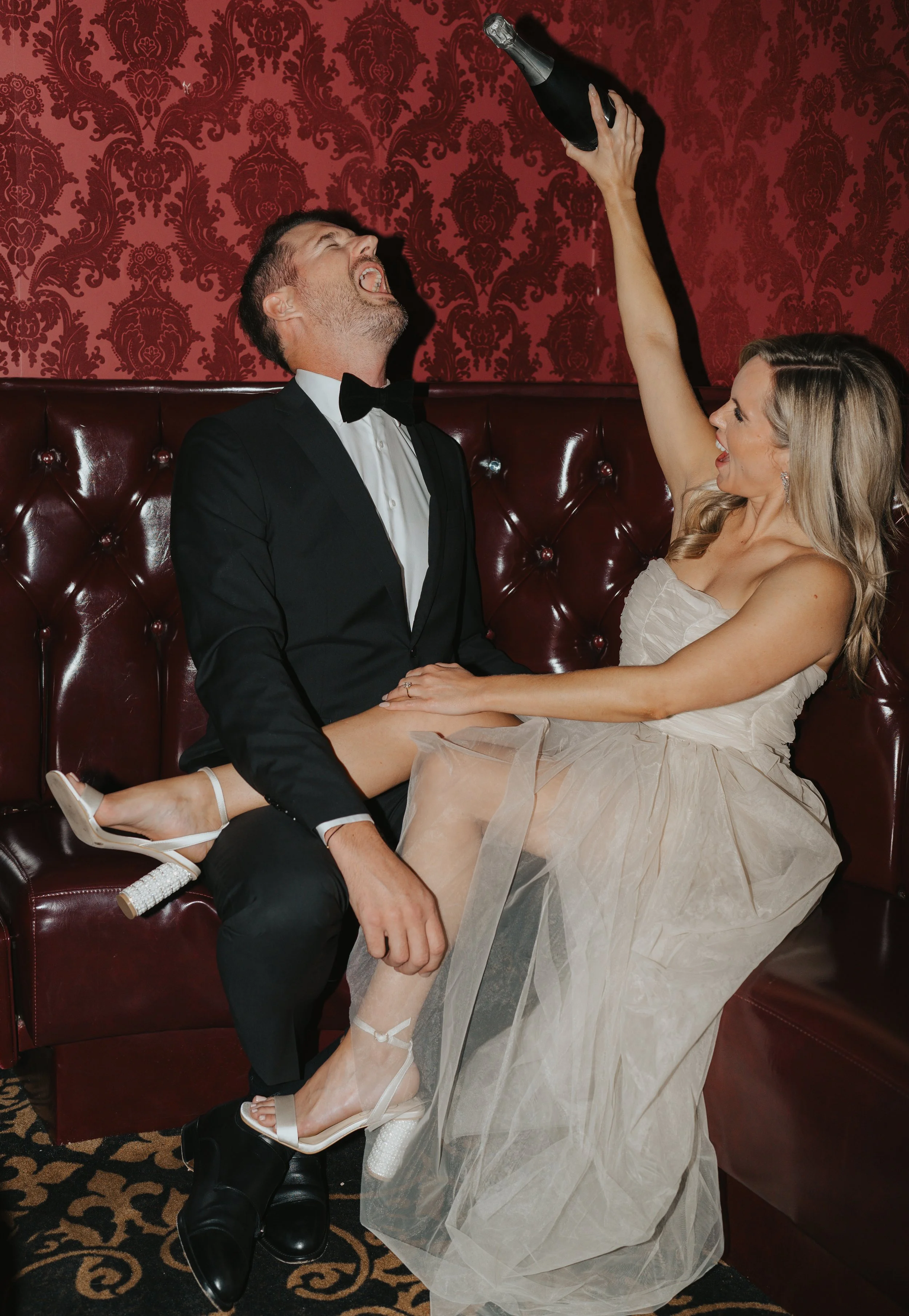 Man and woman celebrating with champagne, woman sitting on man's lap, both dressed in formal attire, in a luxurious setting.