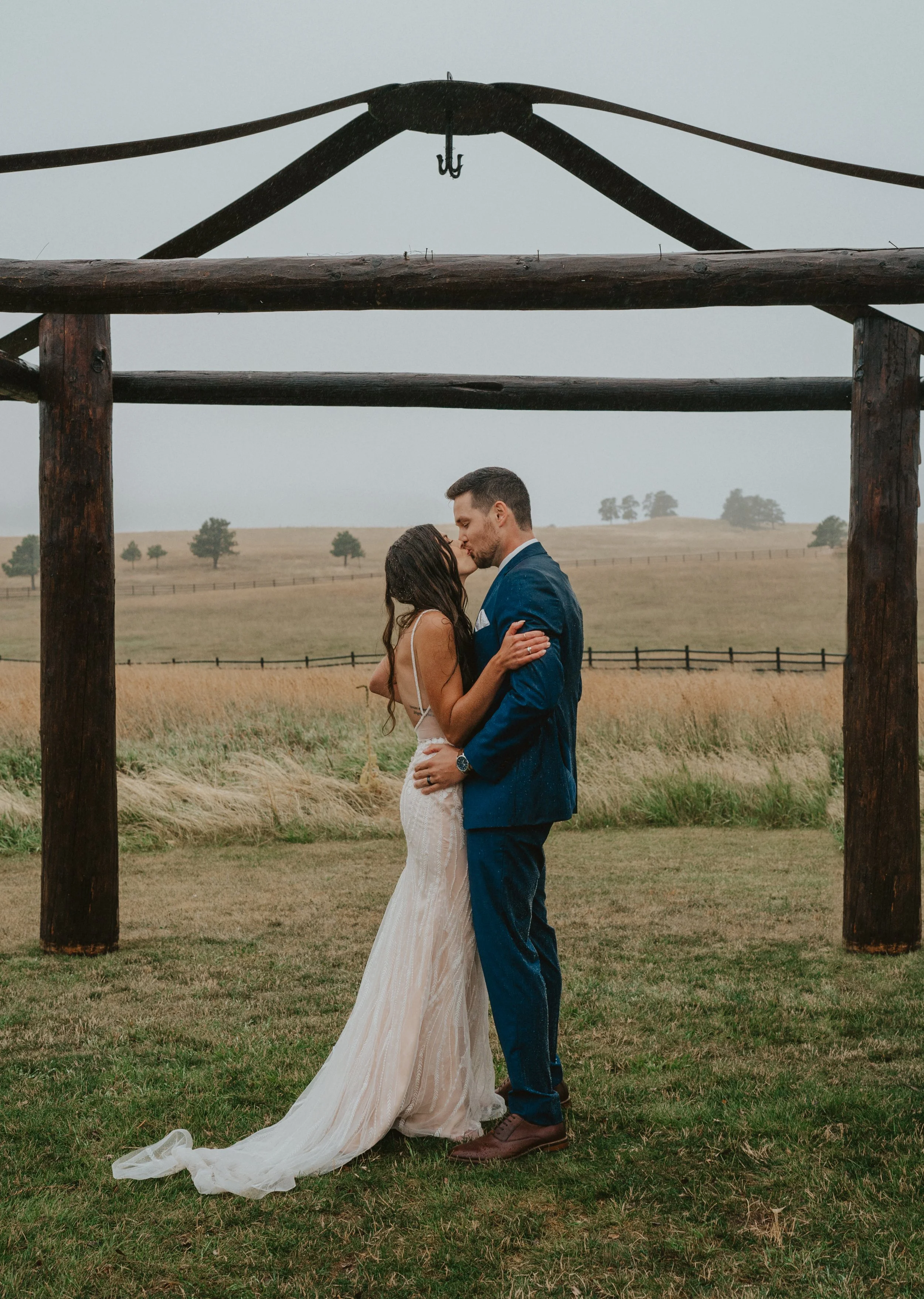 A bride and groom share a kiss under a rustic wooden arch in a field with rolling hills and scattered trees in the background.