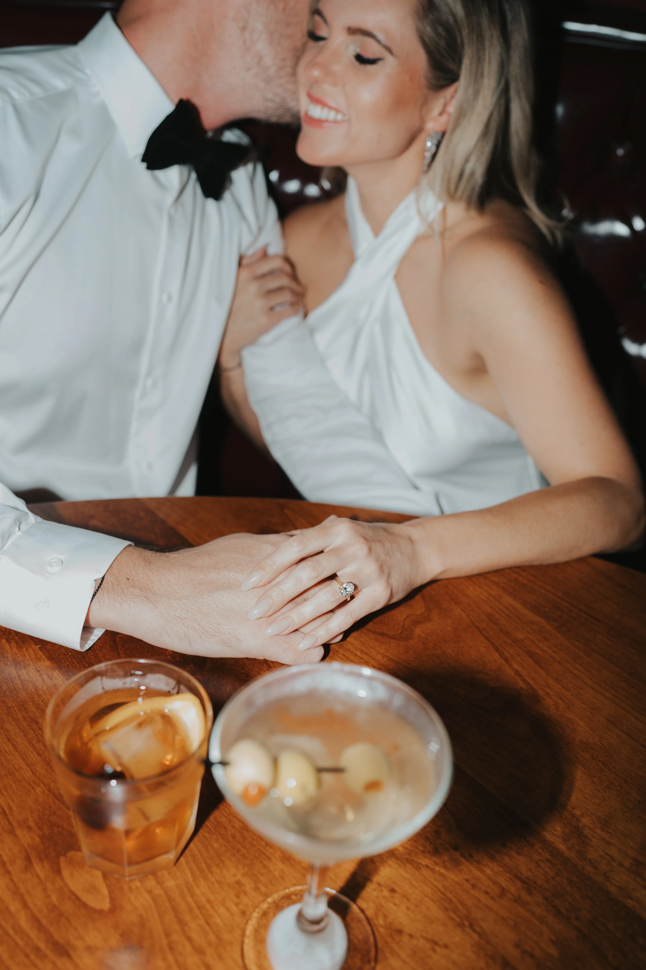 Couple in formal attire sharing a romantic moment at a table, holding hands, with drinks in front of them.