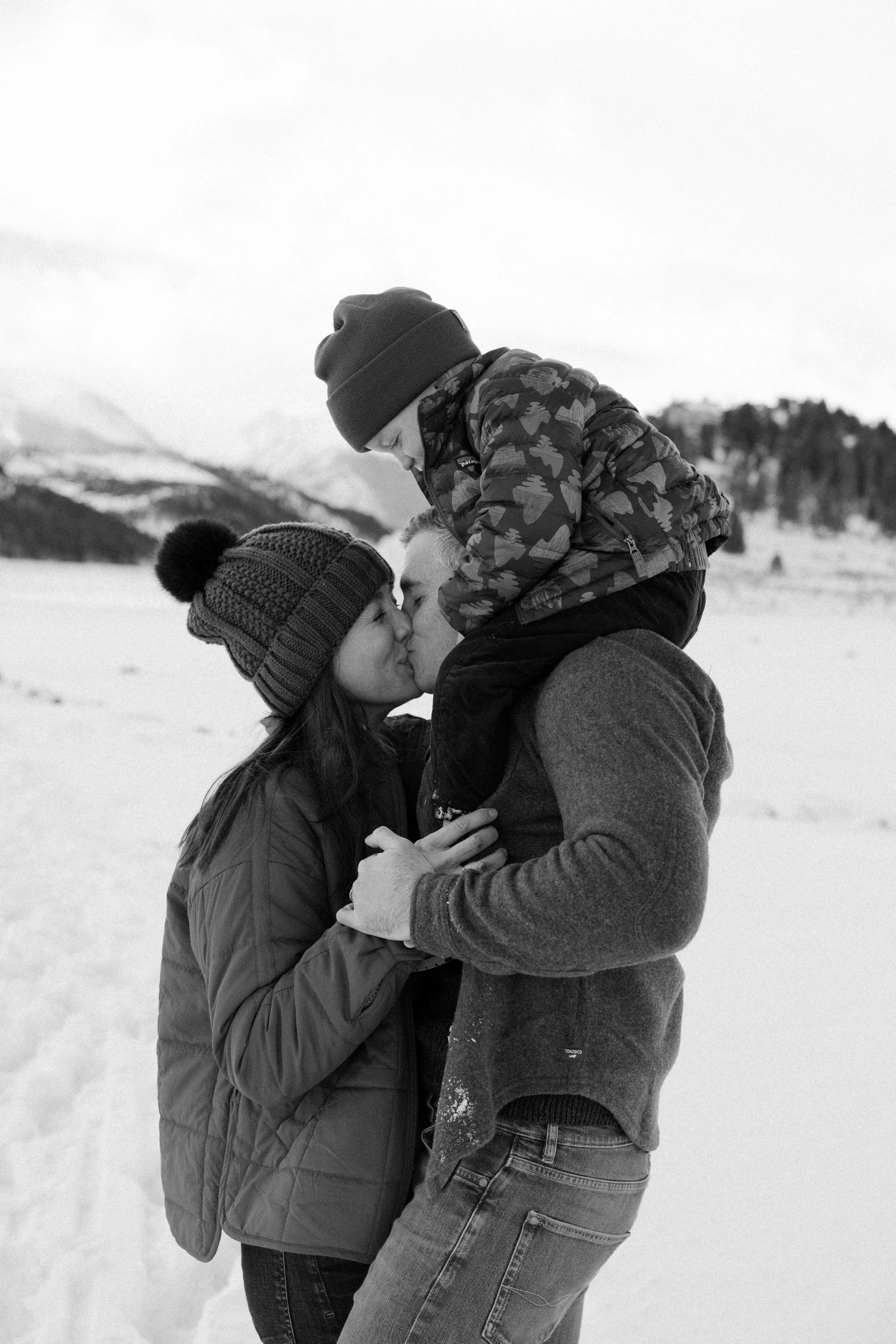 A black and white photo of a family kissing outdoors in a snowy landscape, with mountains in the background.