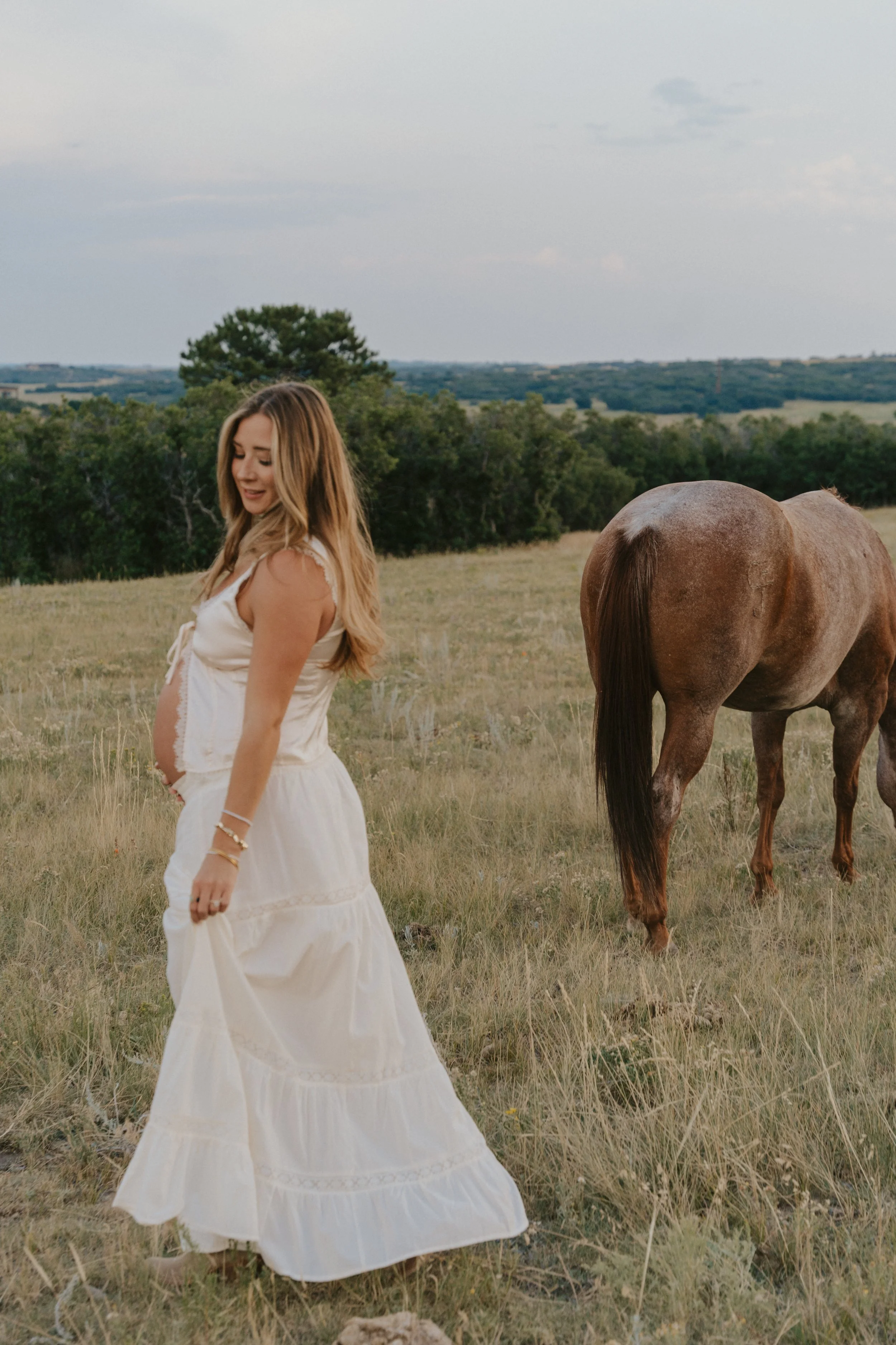 A pregnant woman in a white dress standing in a grassy field near a brown horse during daytime.