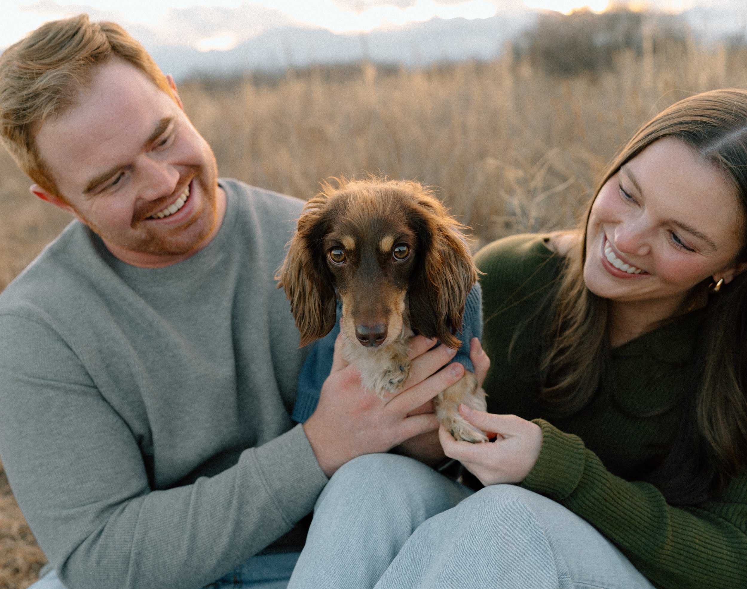 A smiling man and woman sitting outdoors in a field, holding a brown and tan long-haired dachshund between them, with mountains in the background.
