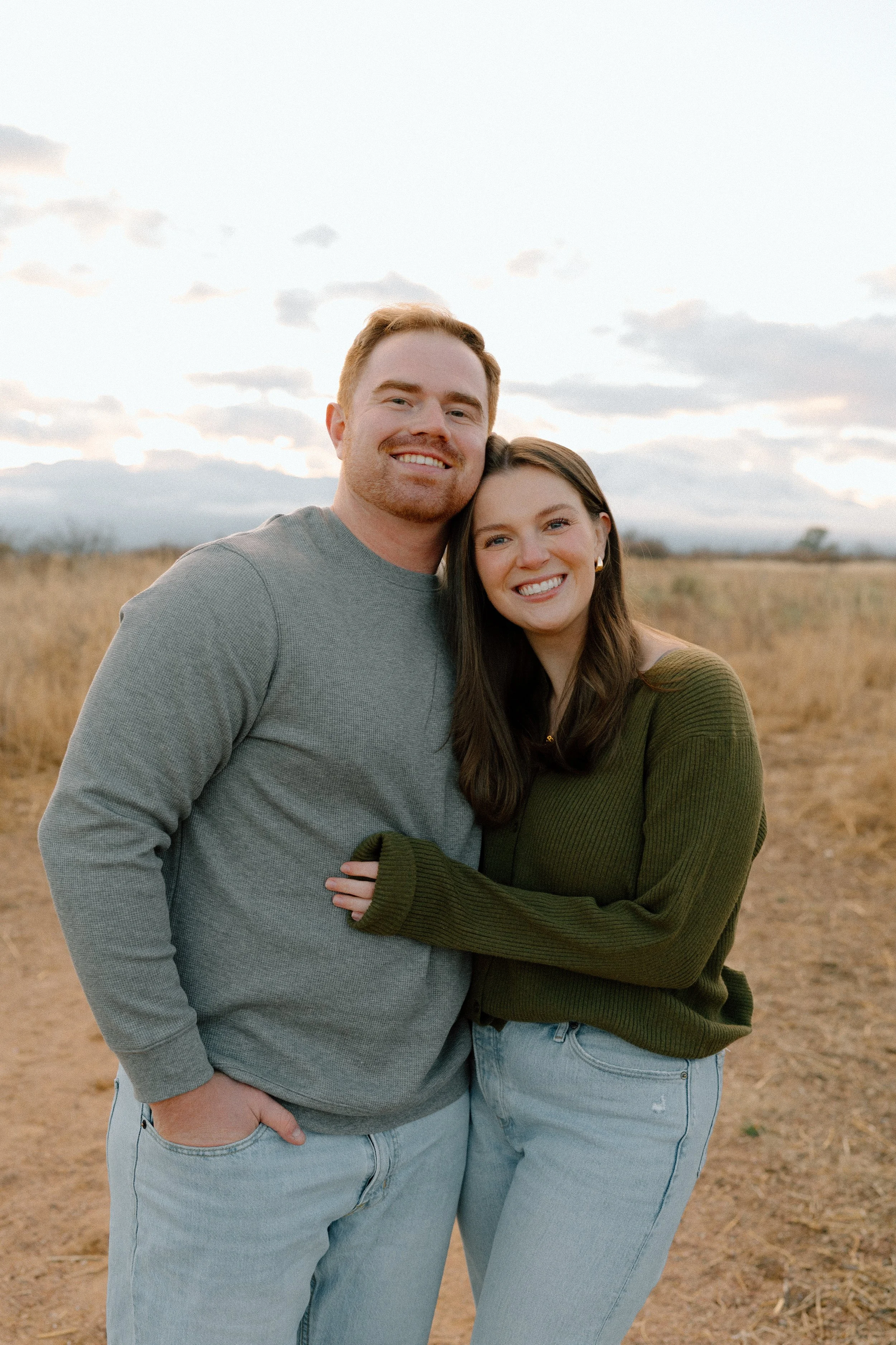 A smiling young couple stands close together outdoors in a field during sunset, with the man wearing a gray long-sleeve shirt and the woman in a green sweater and light jeans.