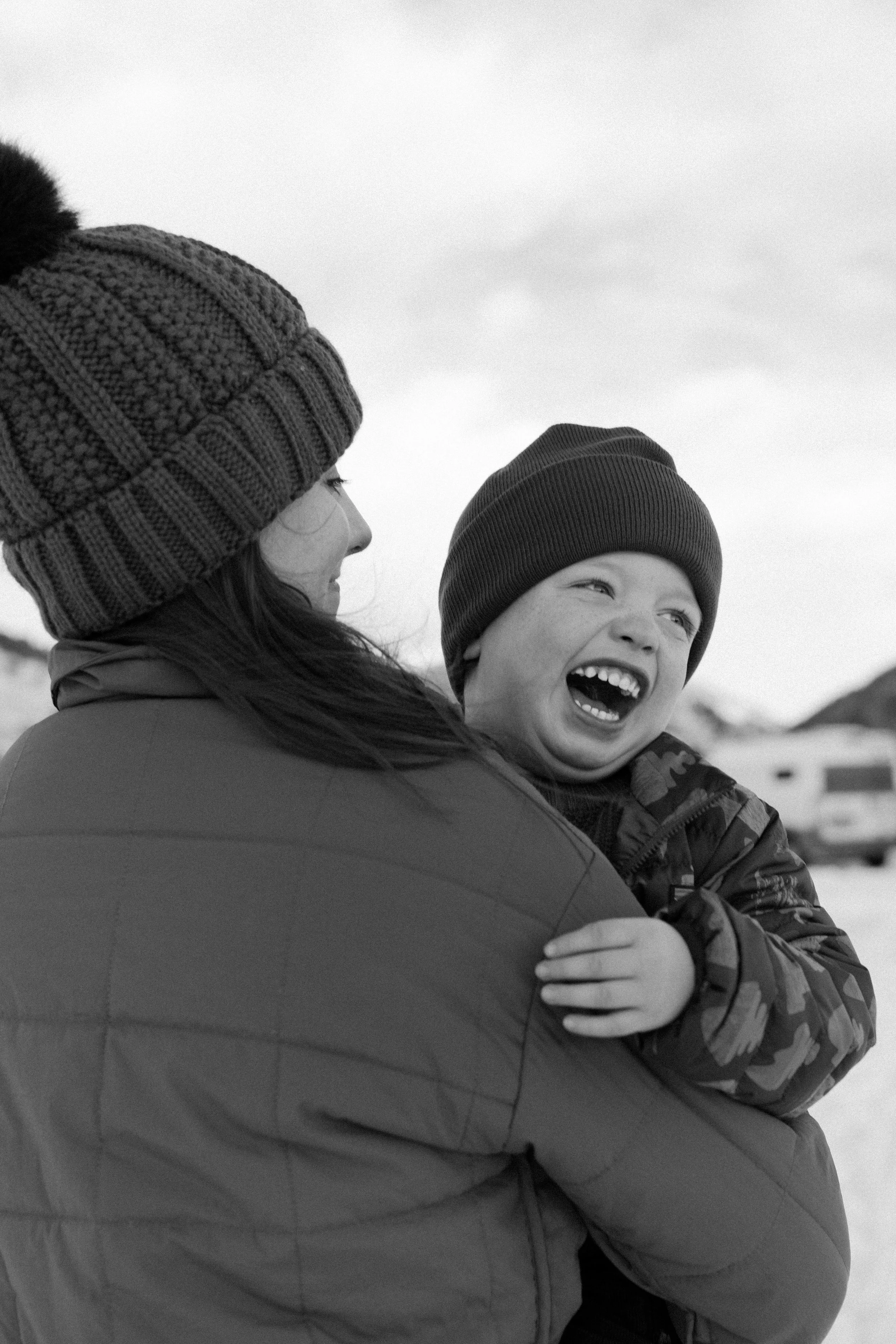 A woman holding a laughing child outdoors in winter, both wearing knit hats and jackets.
