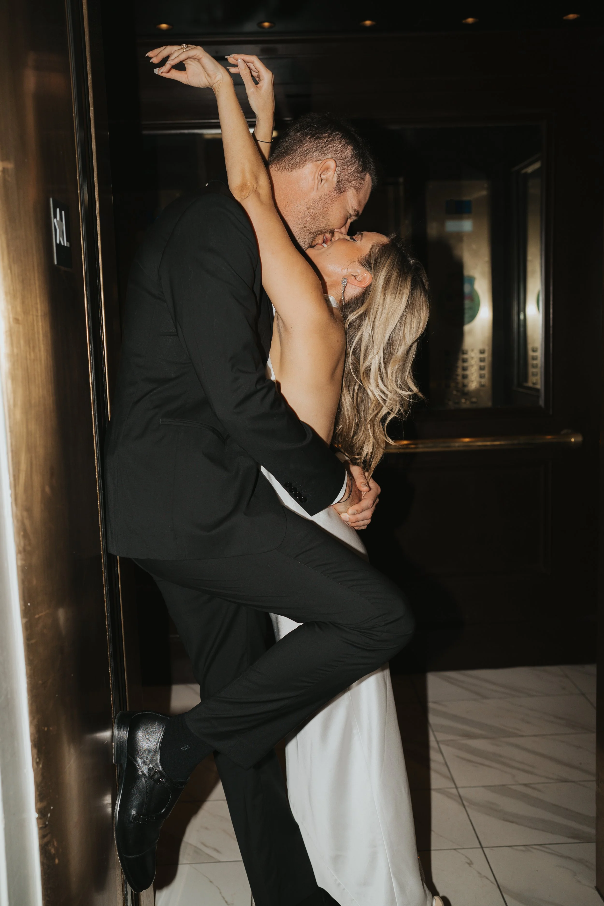 A couple in formal attire sharing a passionate kiss in an elevator, with one person embracing the other.