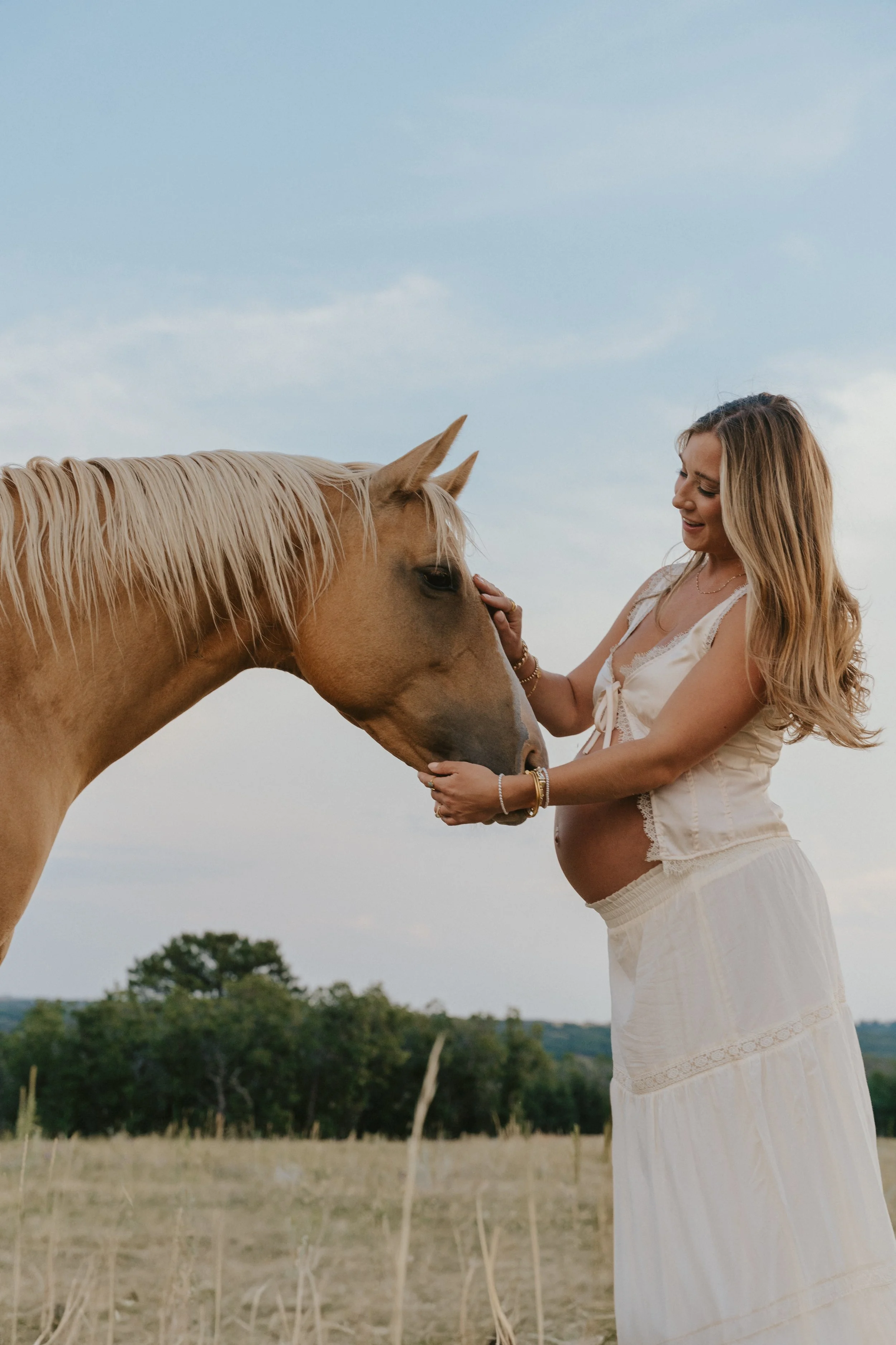 A pregnant woman with long blonde hair wearing a white dress standing outdoors, gently touching the face of a light-colored horse with her hands, smiling at the horse against a blue sky and green trees in the background.