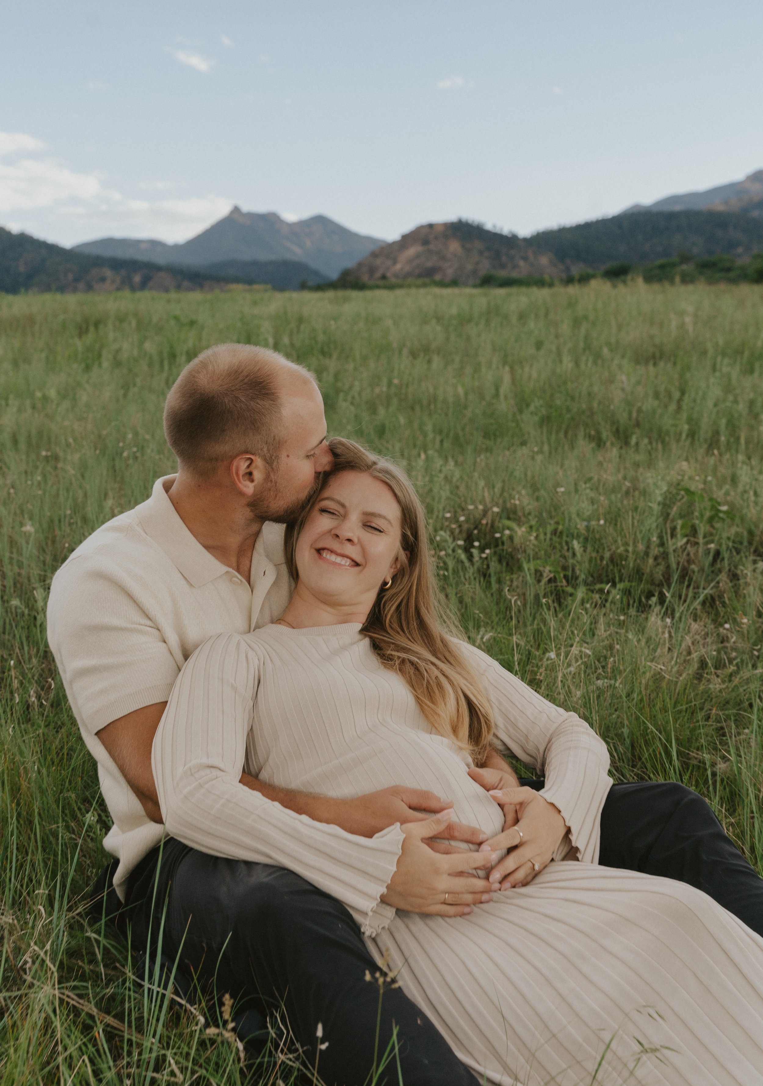 A couple sitting on grass in a field, the man kissing the woman on her forehead, the woman smiling with her eyes closed, in a scenic outdoor setting with mountains in the background.