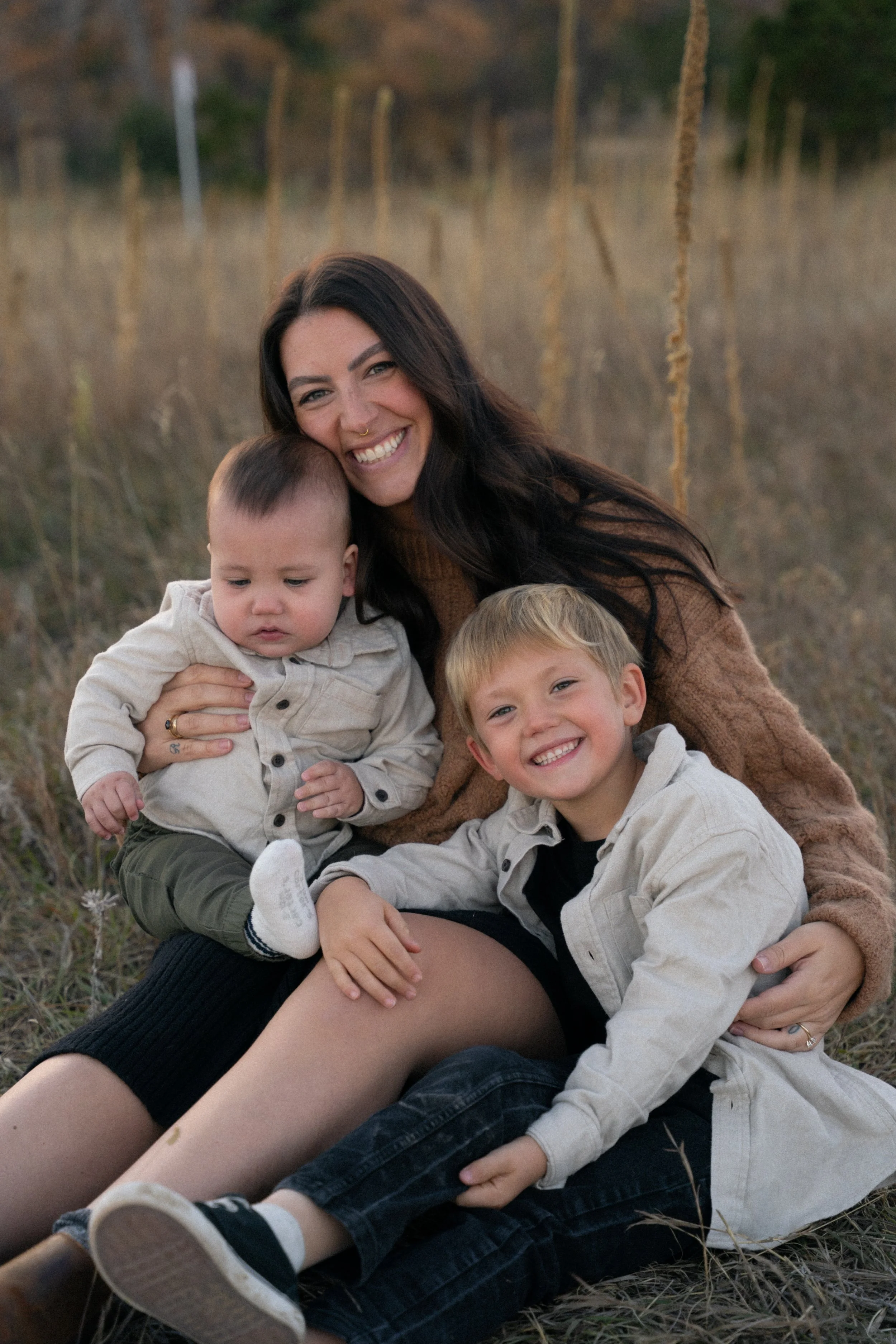 A woman with long dark hair smiling with two young boys sitting on her lap outdoors in a field with tall grass and trees in the background.