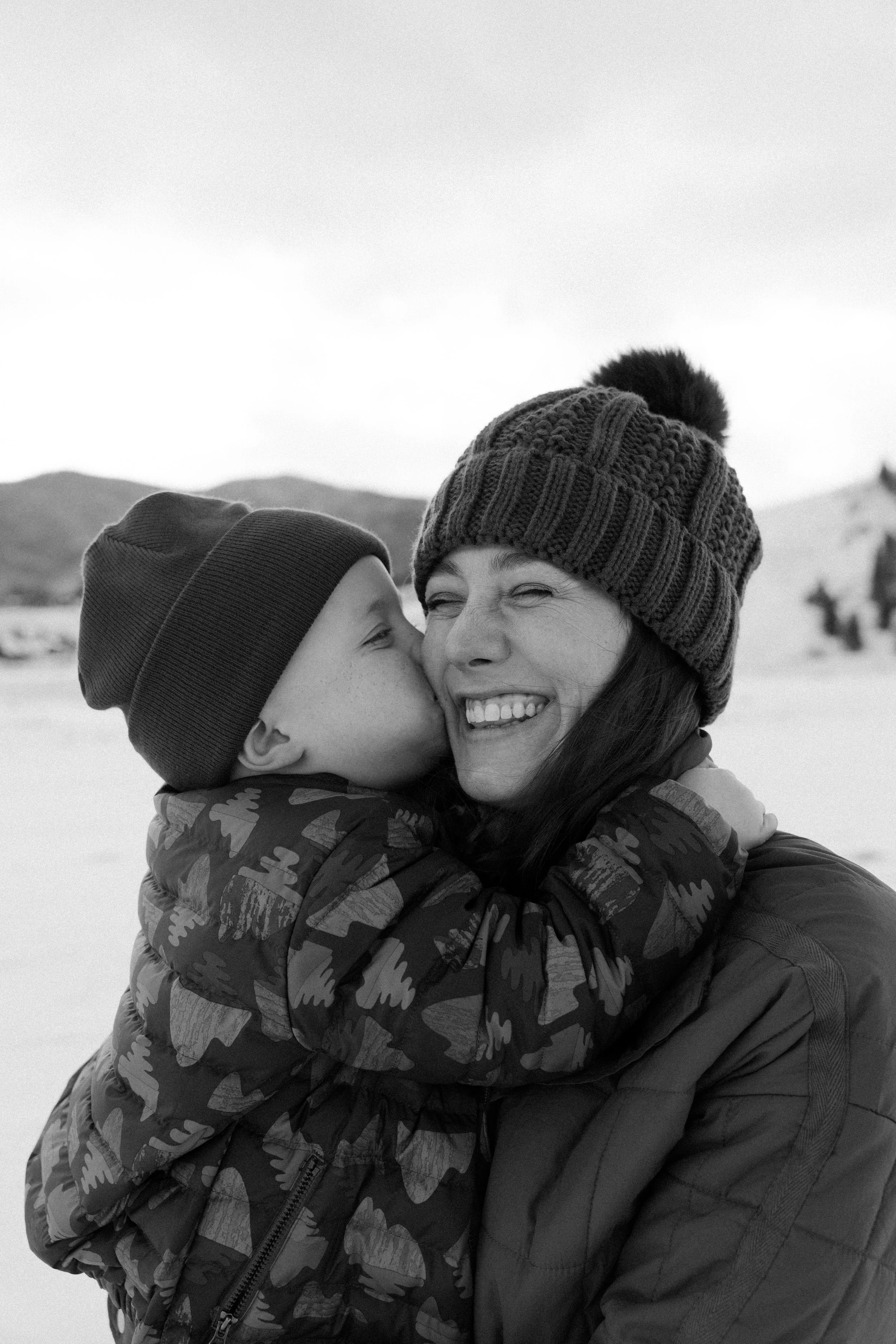 A woman and a young boy share a hug outdoors in a snowy landscape, dressed in winter clothing.