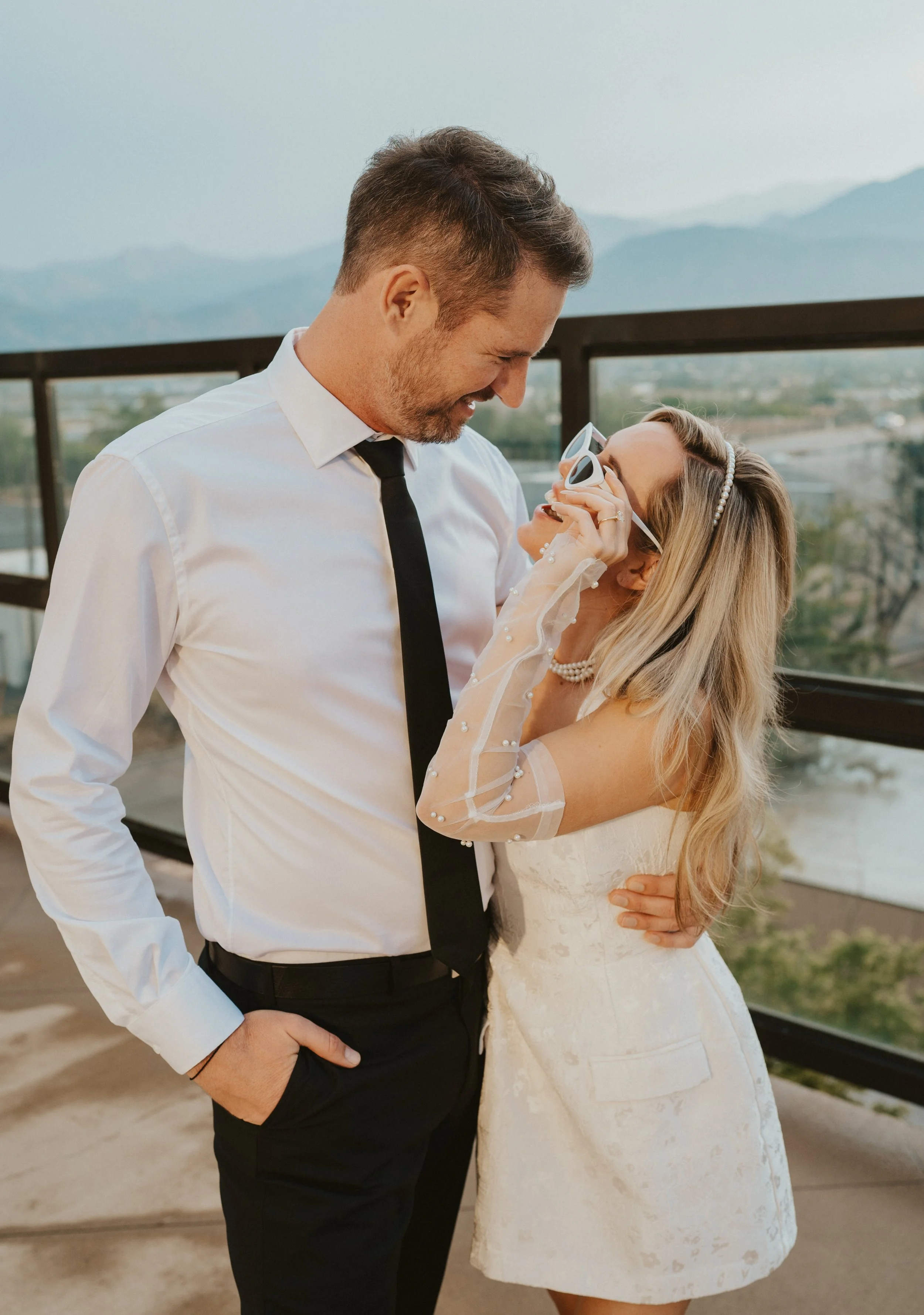 A happy couple on their wedding day, with the woman wearing sunglasses and a white wedding dress, and the man in a white shirt and black tie, sharing a joyful moment on a balcony with scenic mountains and a river in the background.