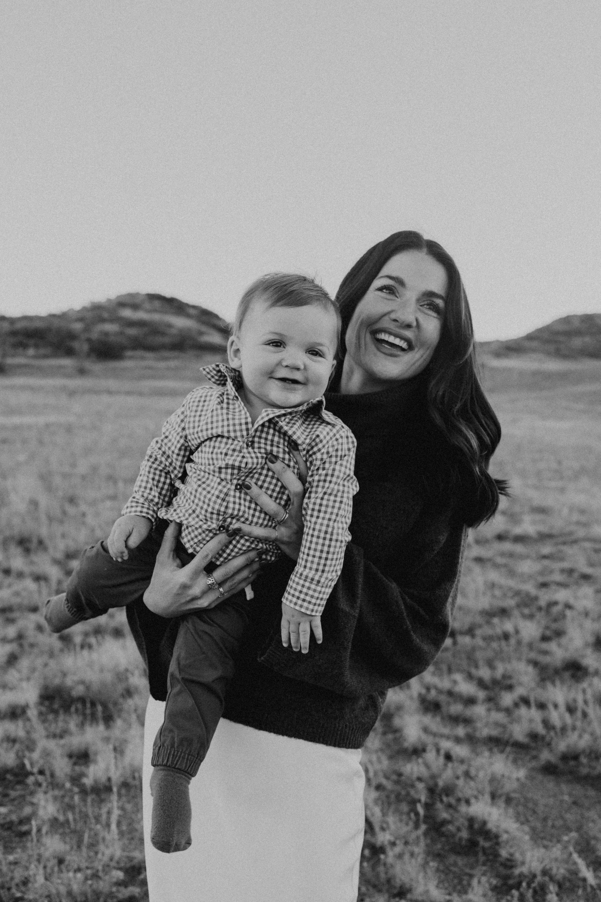 A woman holding a smiling toddler outdoors in a field with hills in the background, in black and white.