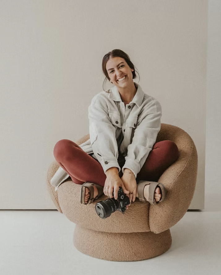 A woman sitting cross-legged on a brown, plush armchair, smiling and holding a camera, in a room with neutral-colored walls.