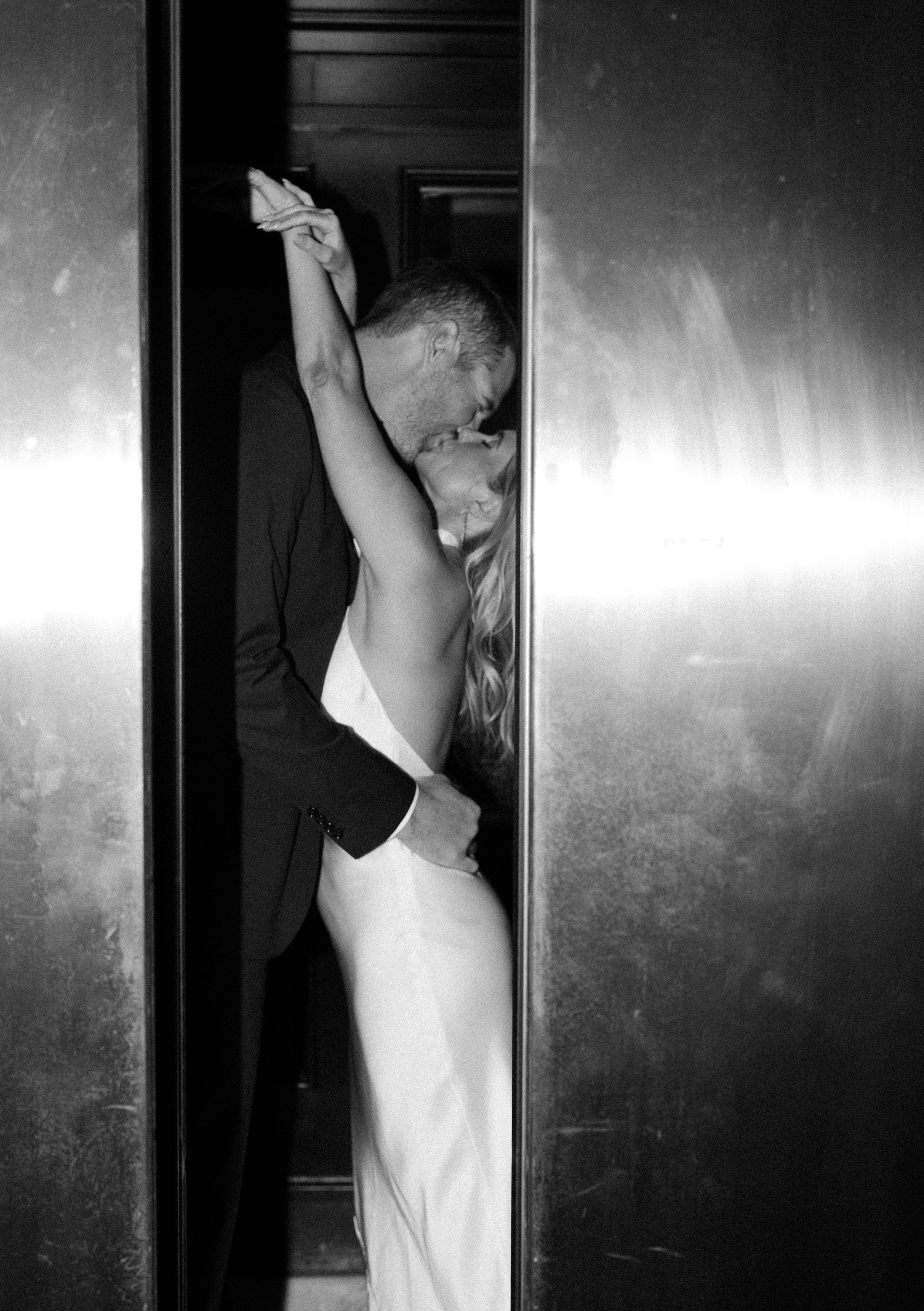 A black and white photo of a couple in an elevator, sharing an intimate kiss. The man is dressed in a suit, and the woman is wearing a white dress with her arms raised around his neck.