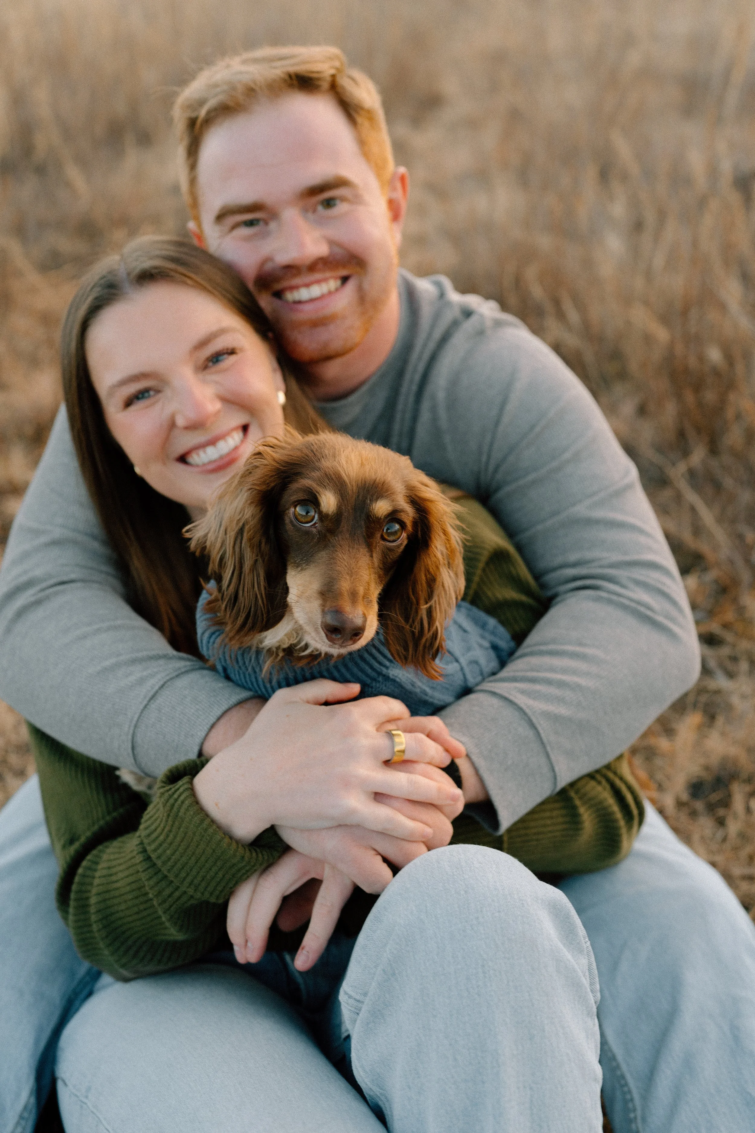A happy couple smiling and hugging with their dog outdoors.