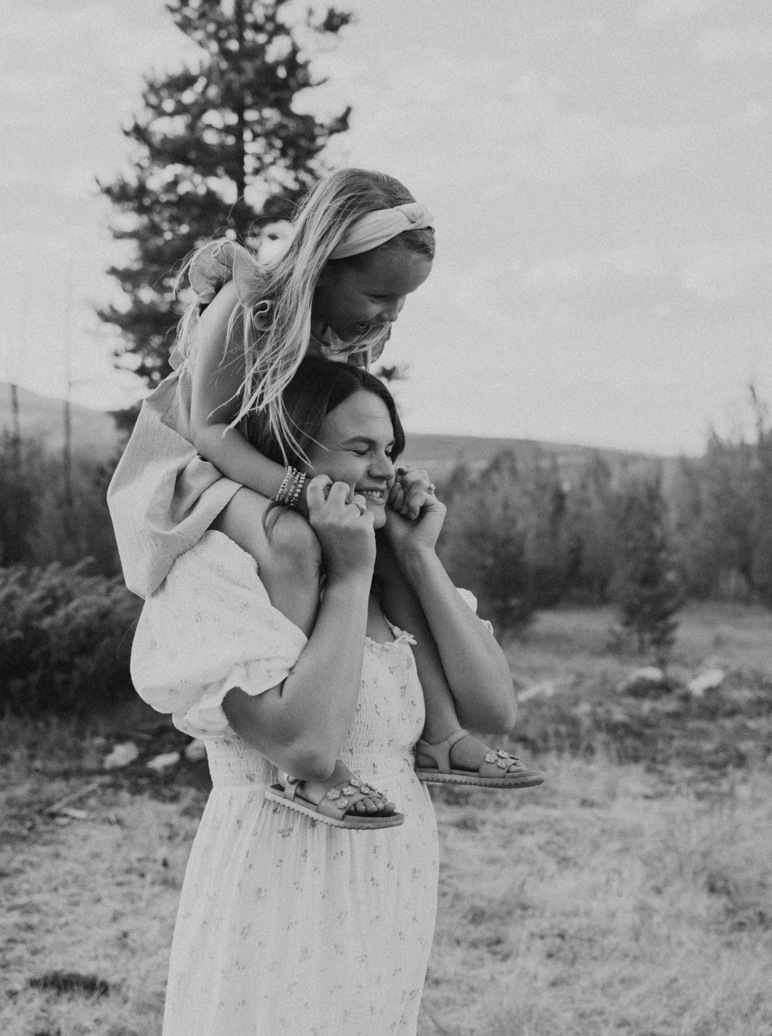 A woman giving a piggyback ride to a young girl outdoors, both smiling and laughing, with trees and a fence in the background.