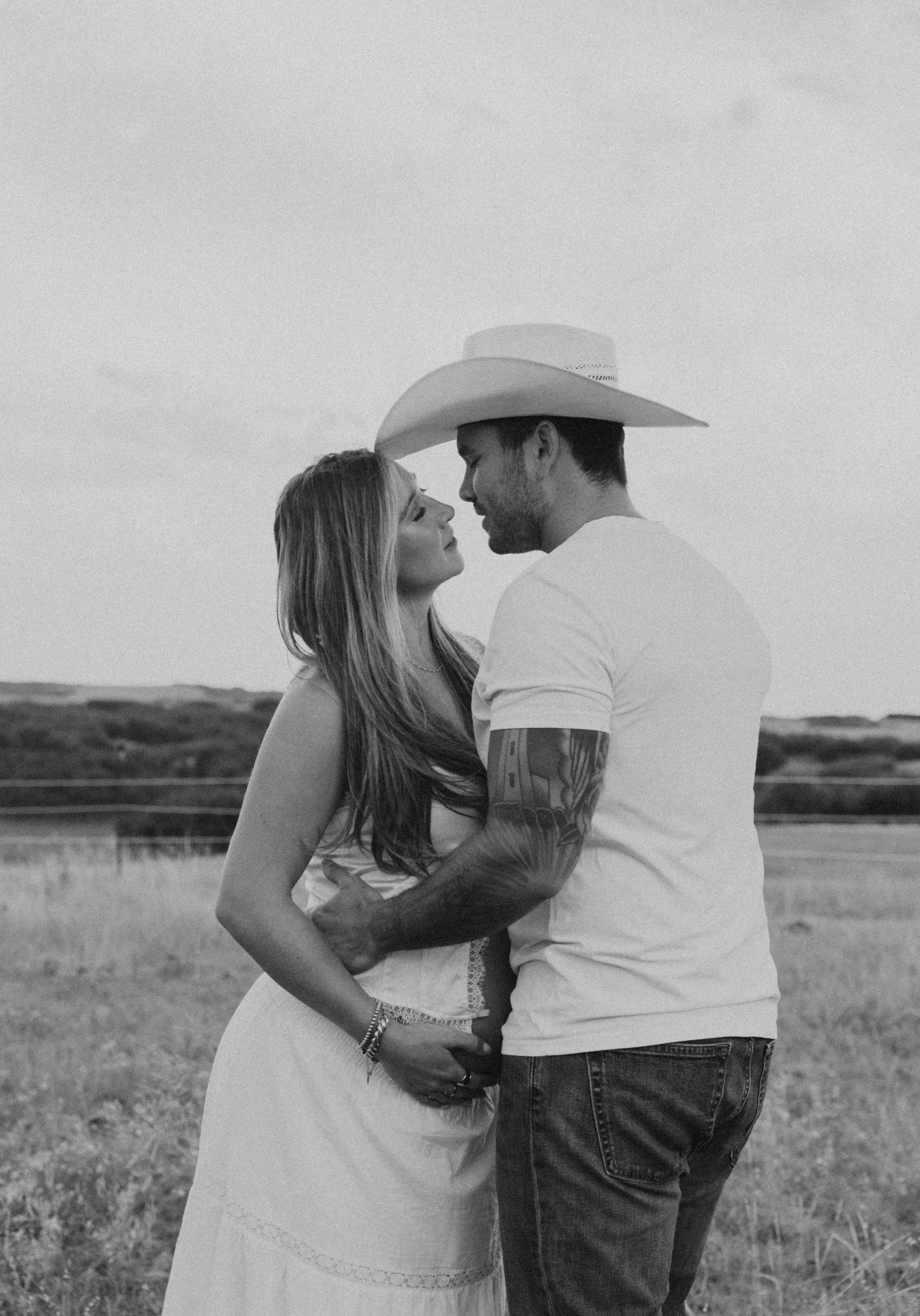 A black and white photo of a couple standing close together outdoors, the woman looking up at the man, who is wearing a cowboy hat, with a background of open fields and a fence.