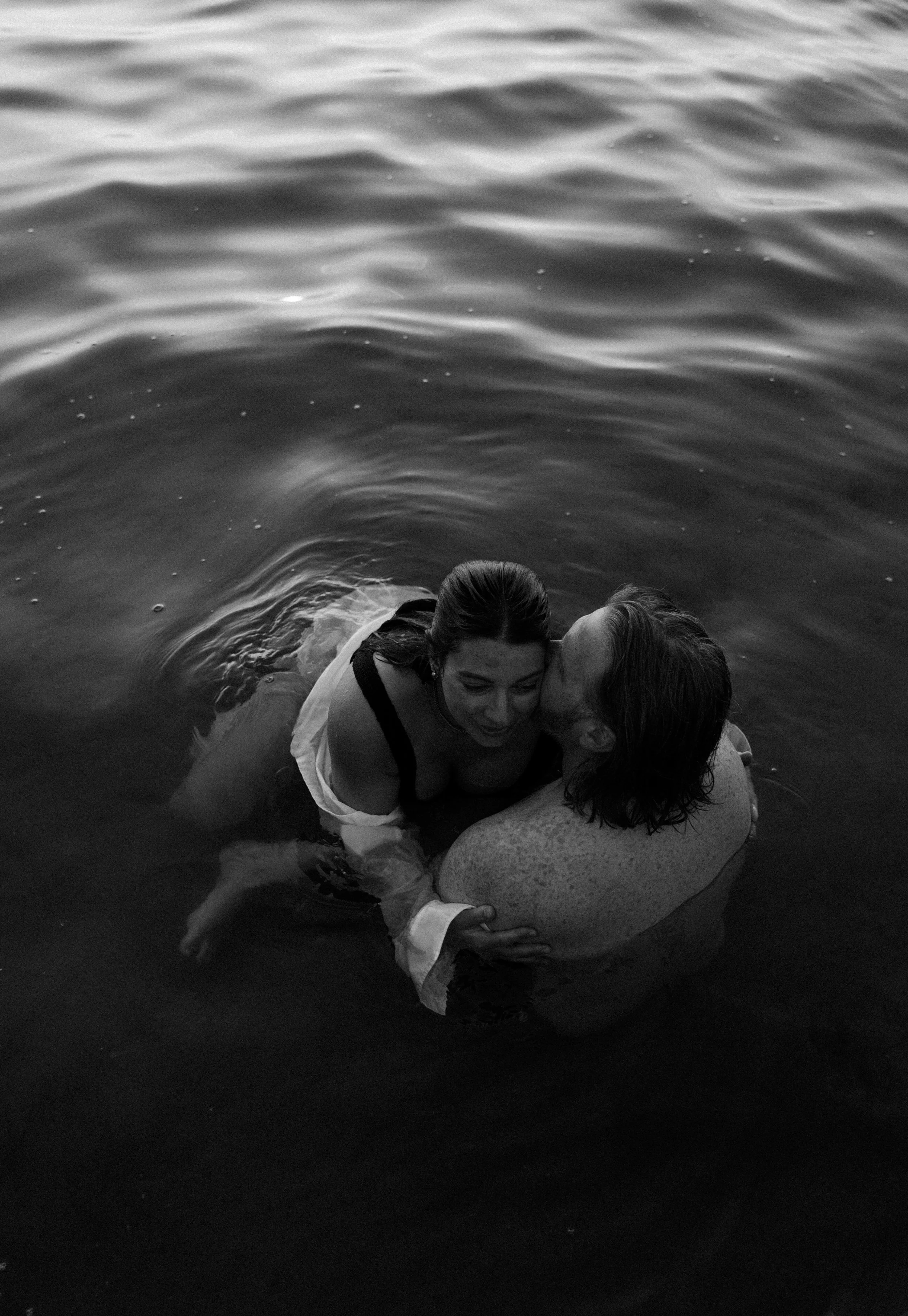 A woman and a man in the water, with the woman kissing the man's cheek. The water around them is calm, and the scene appears to be during sunset or sunrise.