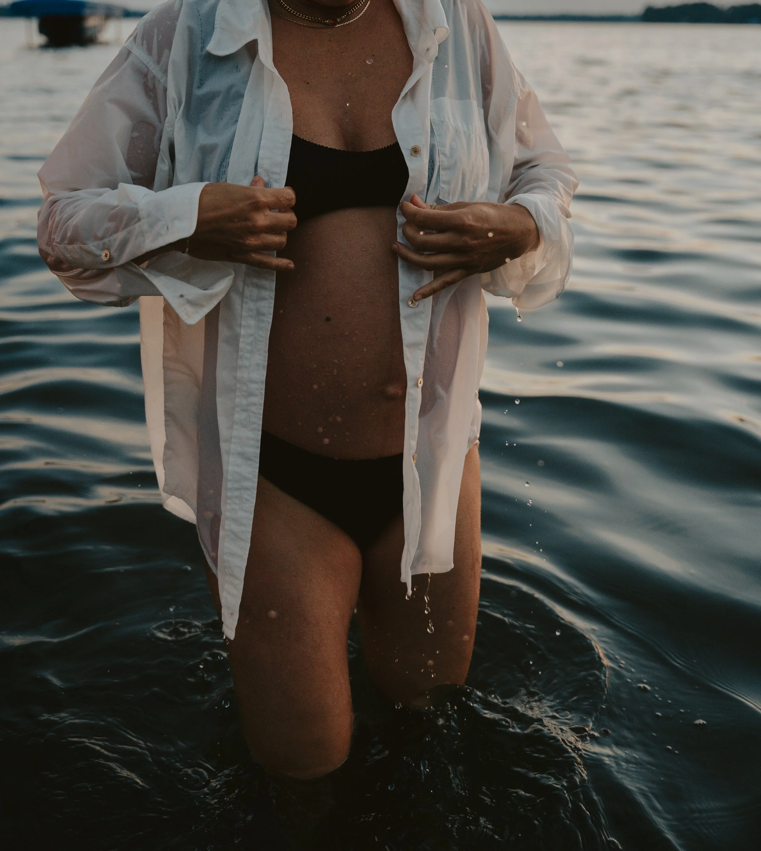 Person standing in water during sunset, wearing a black bikini and a white button-up shirt, with hands near chest and water dripping off body.