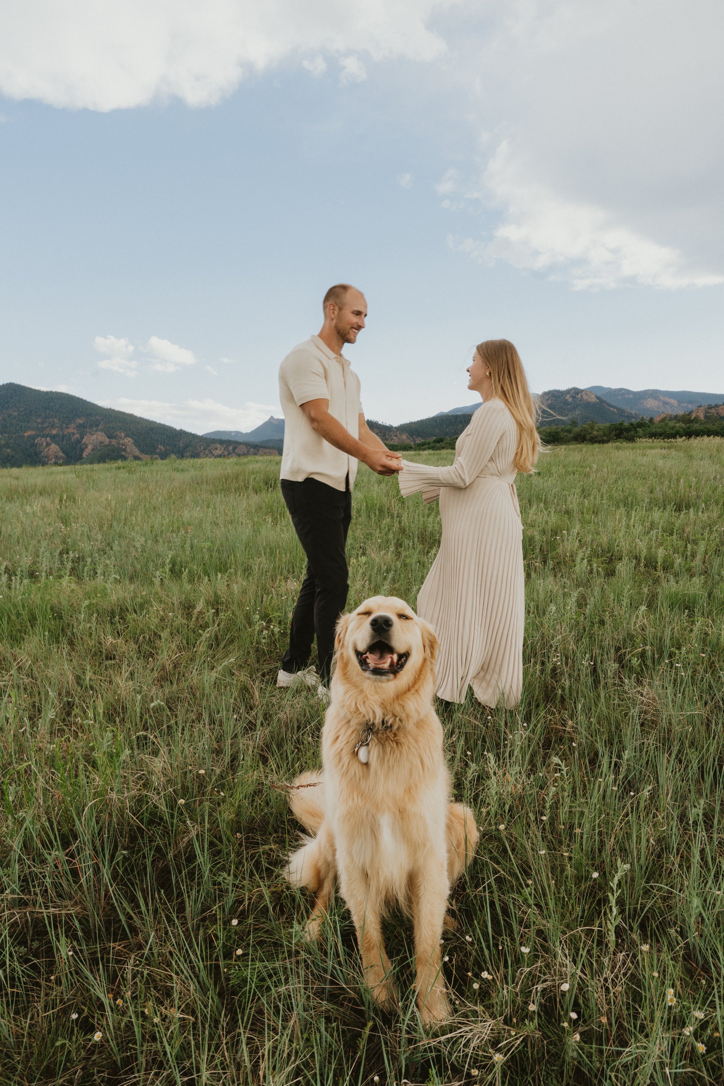 A couple holding hands in a grassy field with mountains in the background, a golden retriever sitting in the foreground, and a blue sky with some clouds.