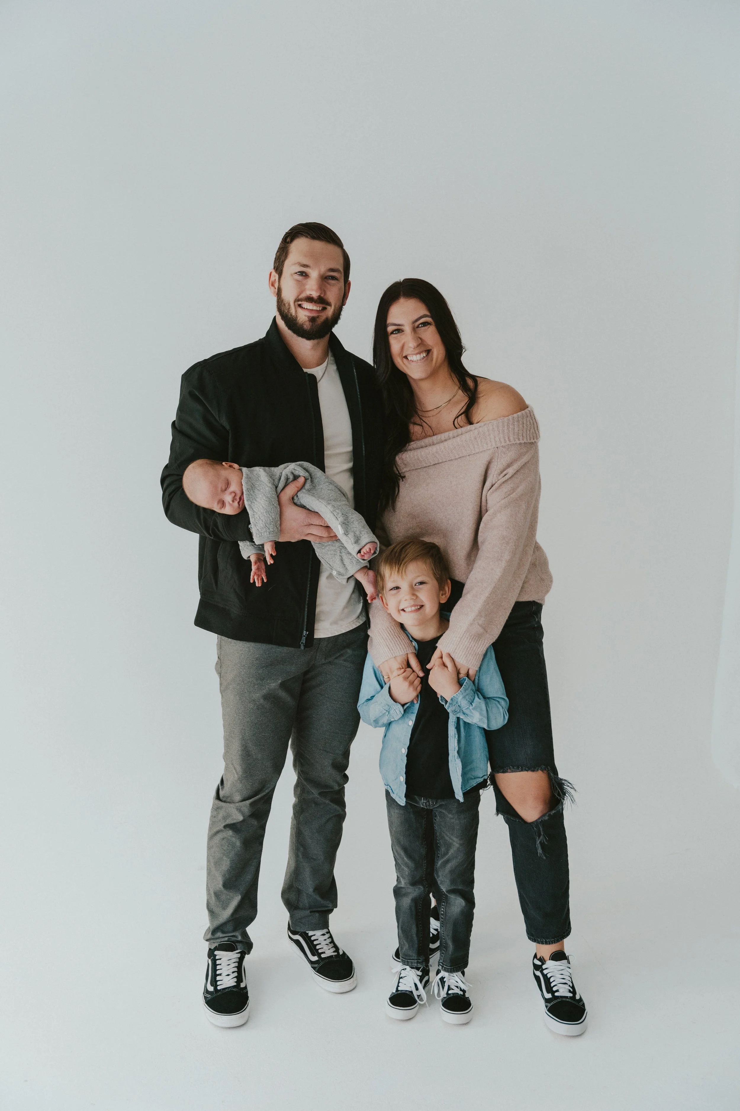 A family of four standing against a plain white background. The father has a beard and is wearing a black jacket, white shirt, and gray pants, holding a sleeping baby. The mother has long black hair, wearing an off-shoulder beige sweater and ripped jeans. The young boy is in front, smiling, with a denim jacket and black shirt. They are all happy and smiling.