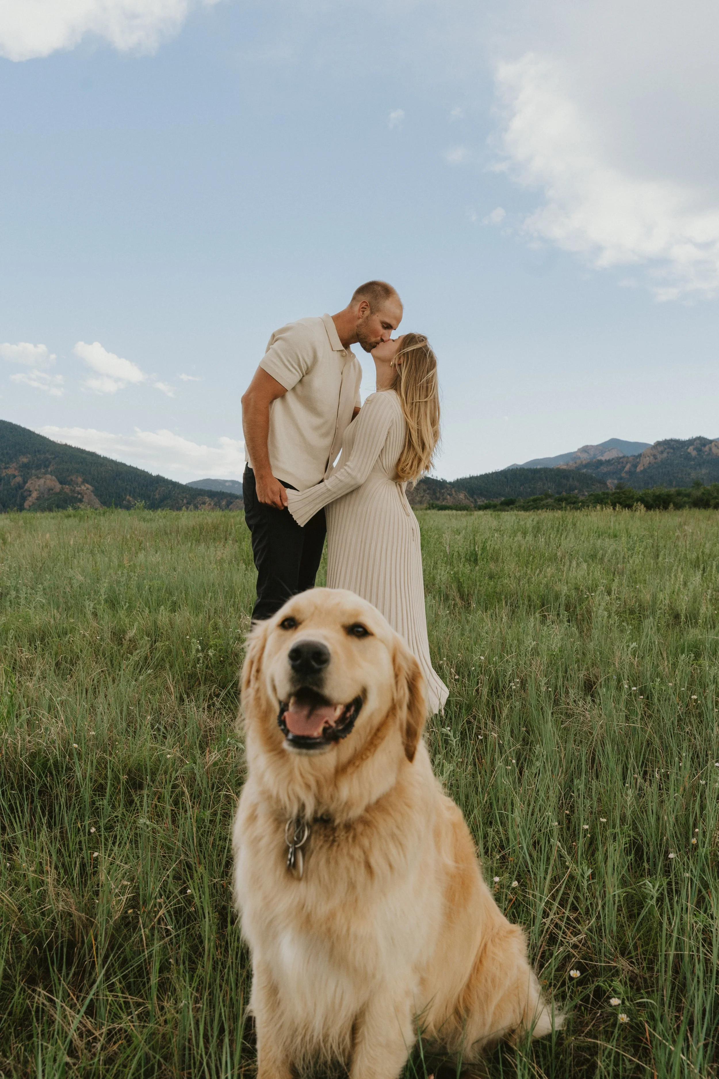 A couple sharing a kiss in a grassy field with mountains in the background, and a golden retriever sitting in the foreground.