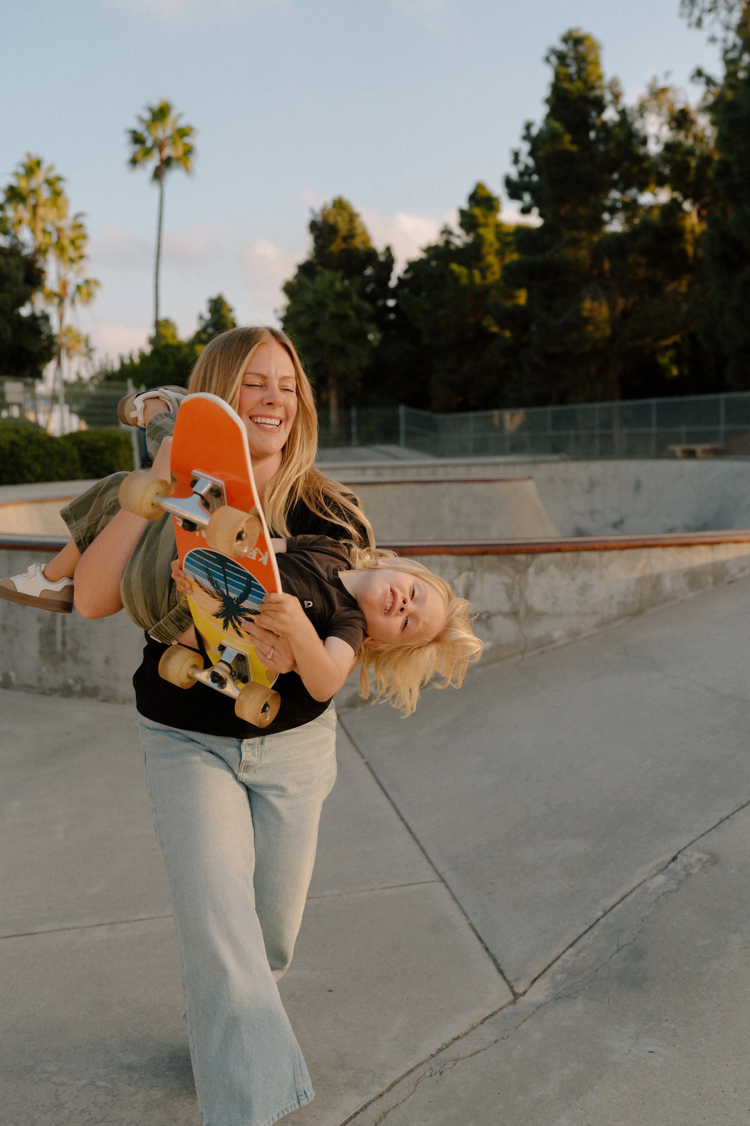 A woman carrying a young girl with a skateboard at a skate park, smiling and laughing as they enjoy their time outdoors during sunset.