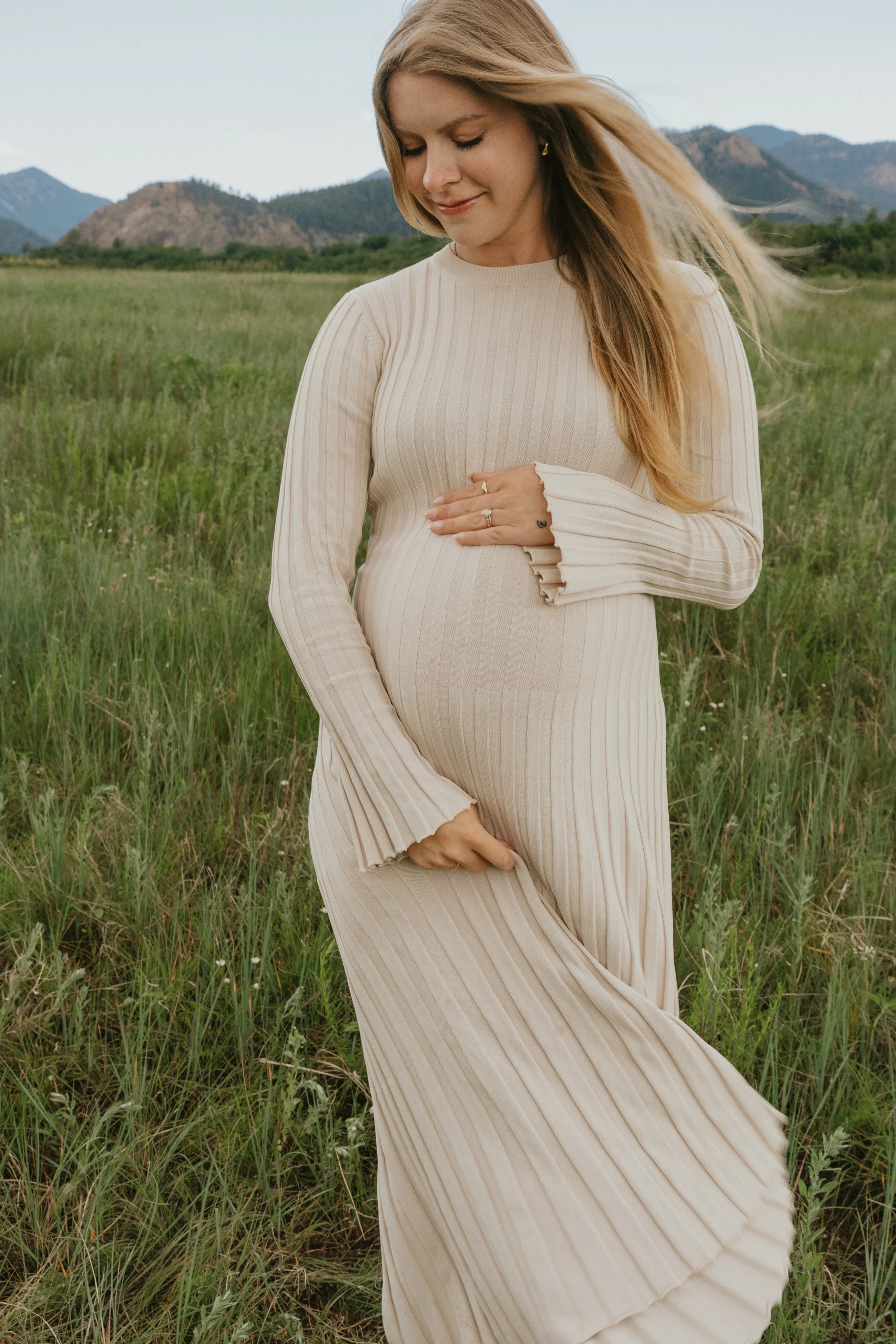 A pregnant woman in a beige ribbed dress standing in a grassy field, gently touching her belly, with mountains in the background and her hair flowing in the wind.