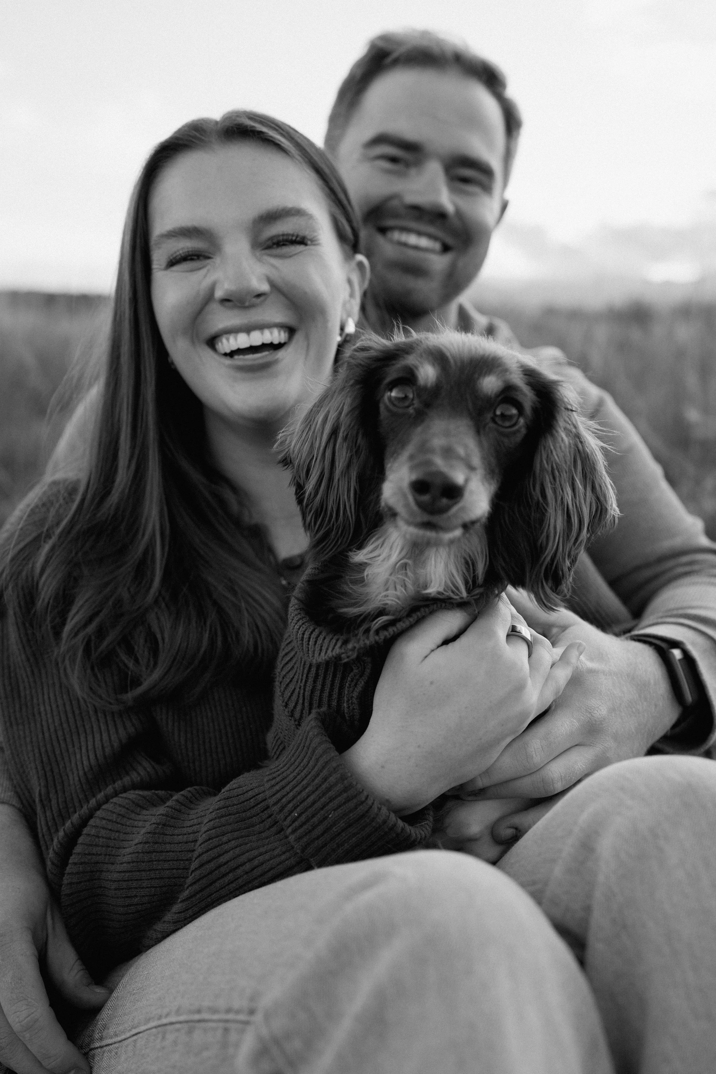 Smiling woman holding a dog with a smiling man in the background outdoors.
