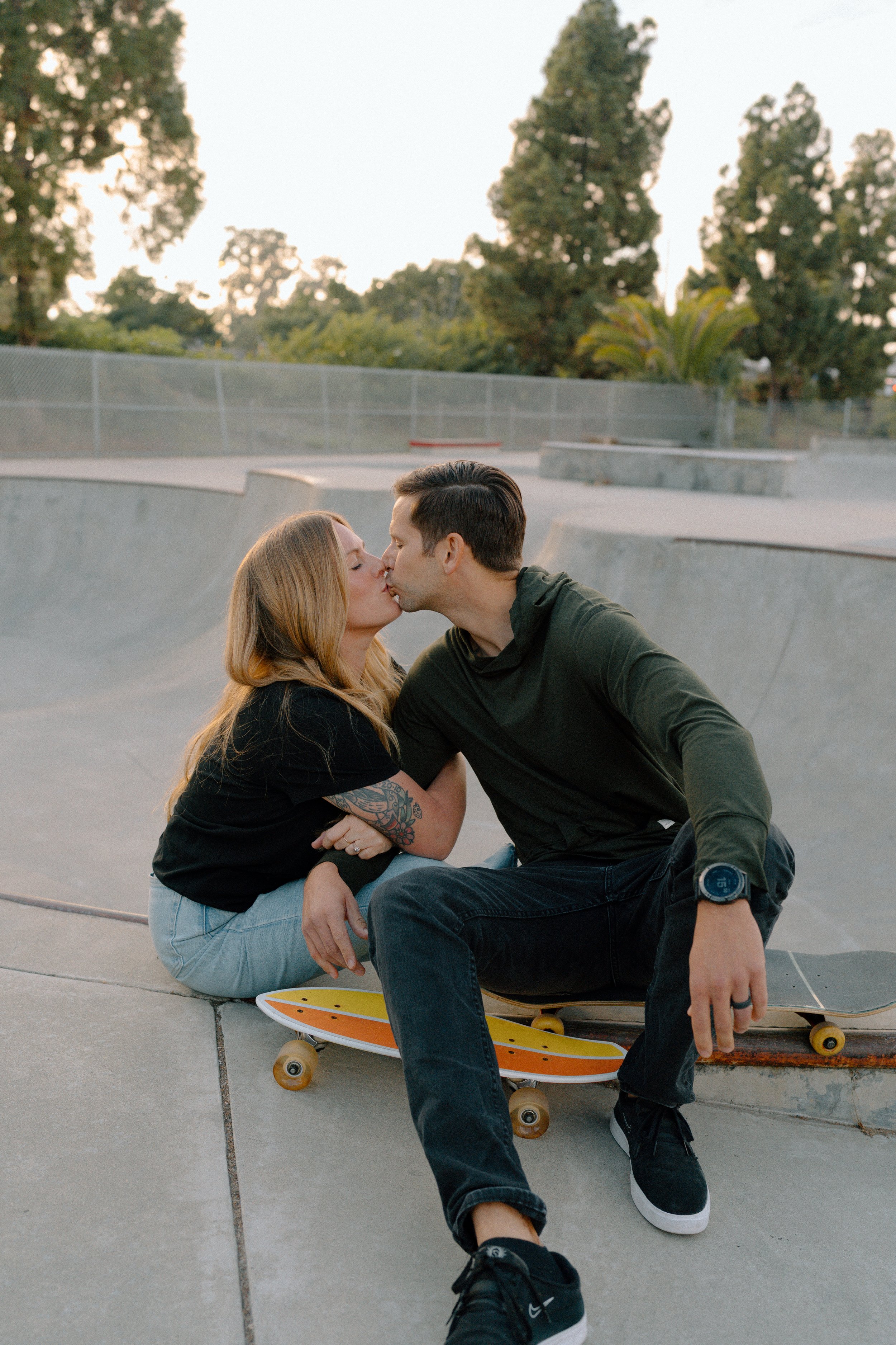 A couple sharing a kiss at a skate park, sitting on the concrete ledge with skateboards nearby, surrounded by trees and an empty skate bowl.