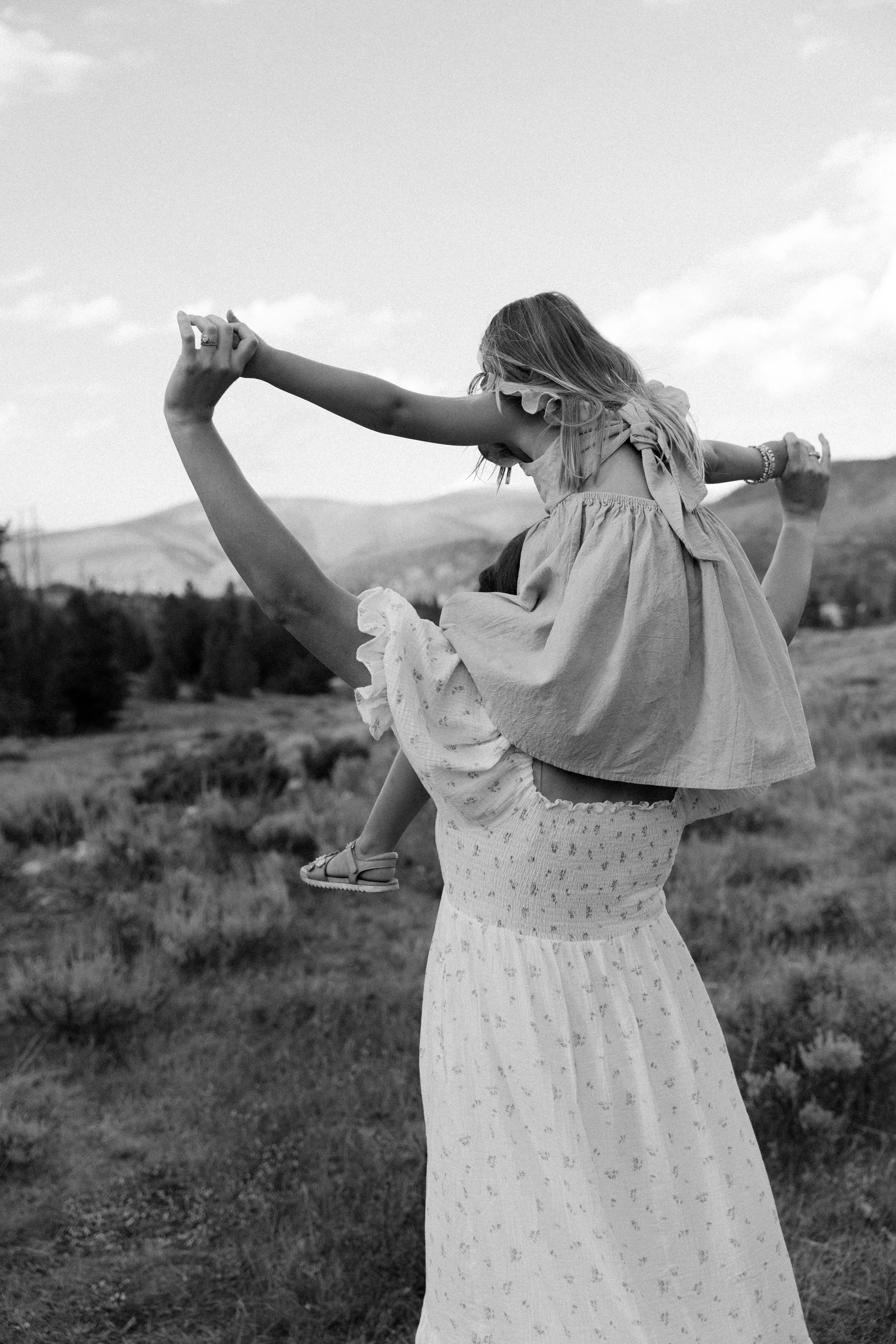 A woman lifting a young girl on her shoulders outdoors in a grassy field, with mountains and cloudy sky in the background. The image is in black and white.