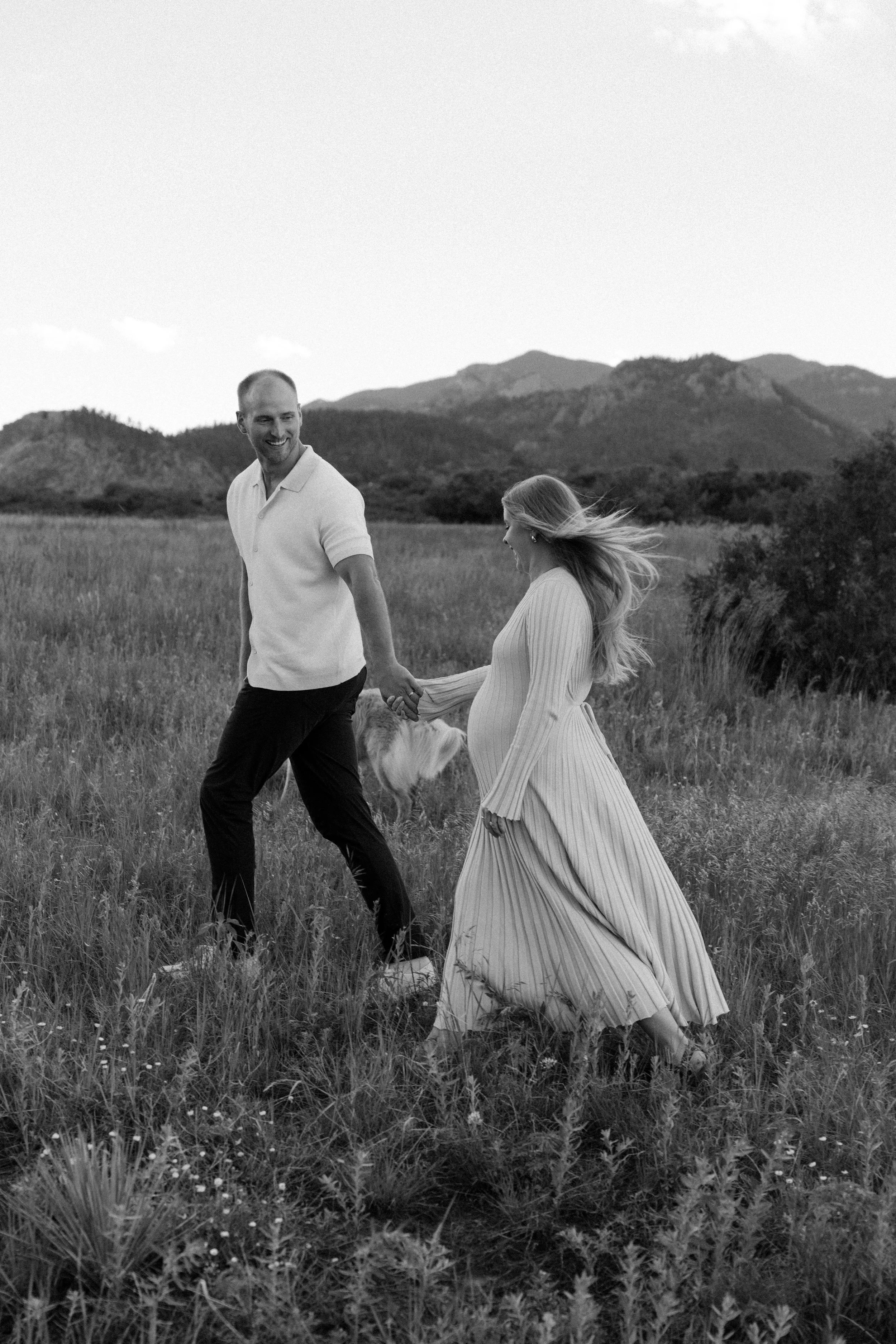 Black and white photo of a couple walking hand in hand through a grassy field, holding a dog. The woman is pregnant, wearing a long, flowing, striped dress, and the man is in a polo shirt and dark pants. Mountains are in the background.