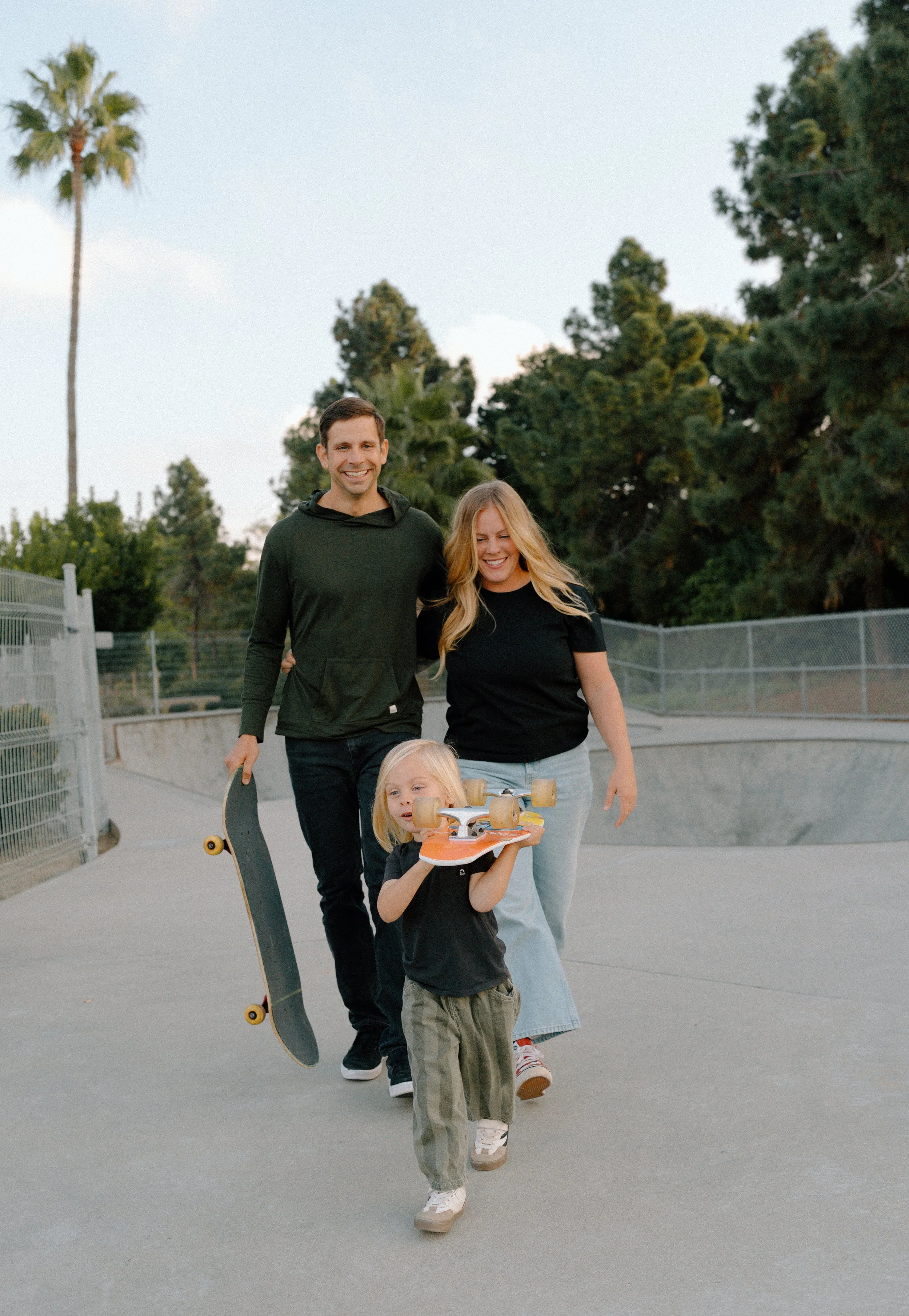 A family of four walking together at a skate park, with the father holding a skateboard and the daughter carrying a skateboard, all smiling and enjoying the outdoors.