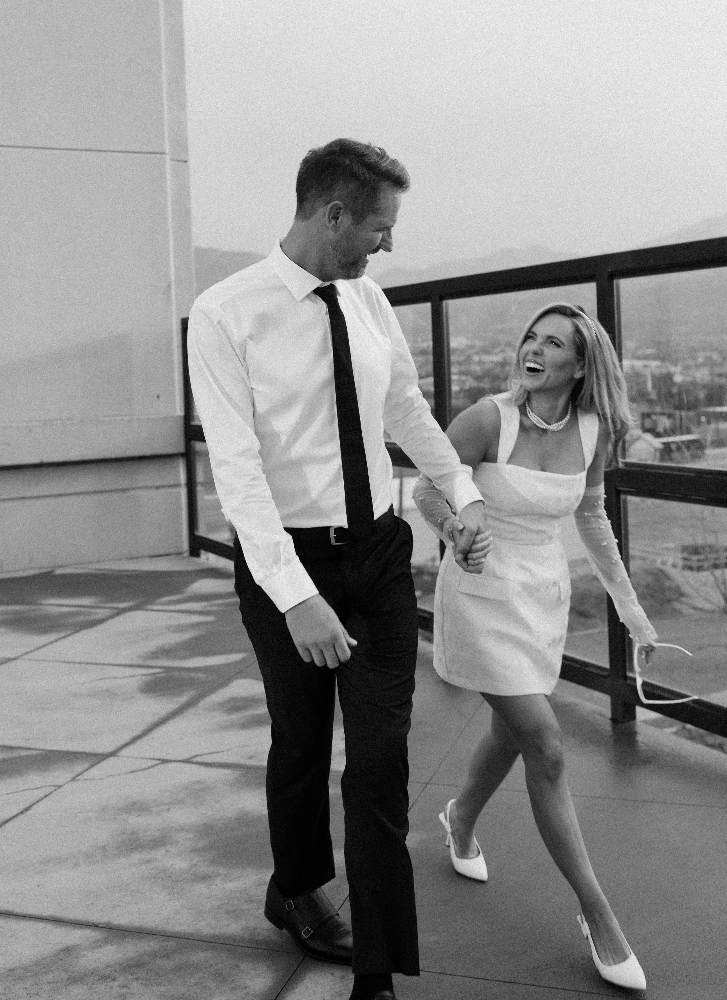 A black-and-white photo of a man and woman holding hands and smiling on a rooftop, with cityscape in the background.