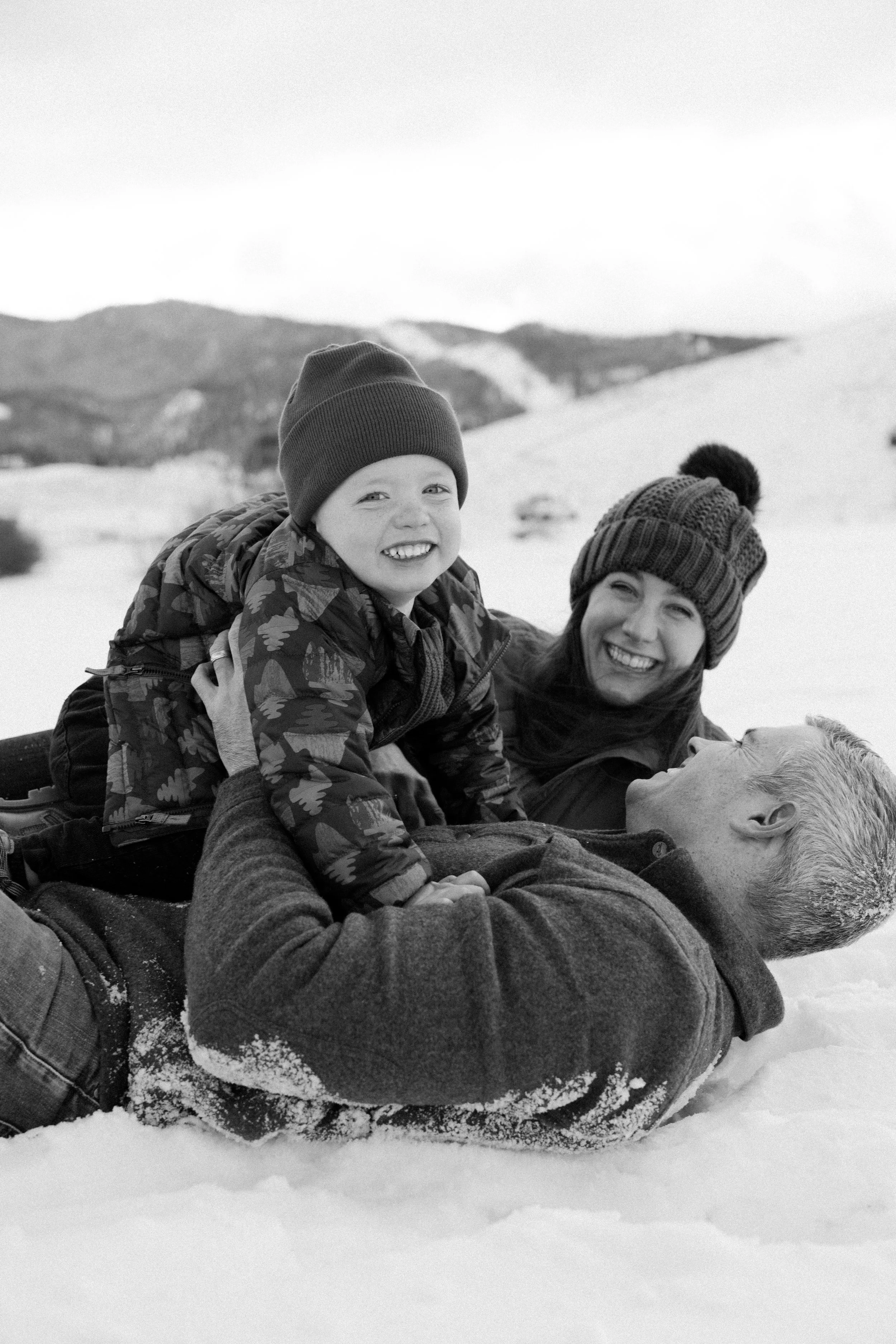 A family of three, including a man, a woman, and a young boy, playing and laying in the snow outdoors during winter, with mountains in the background.