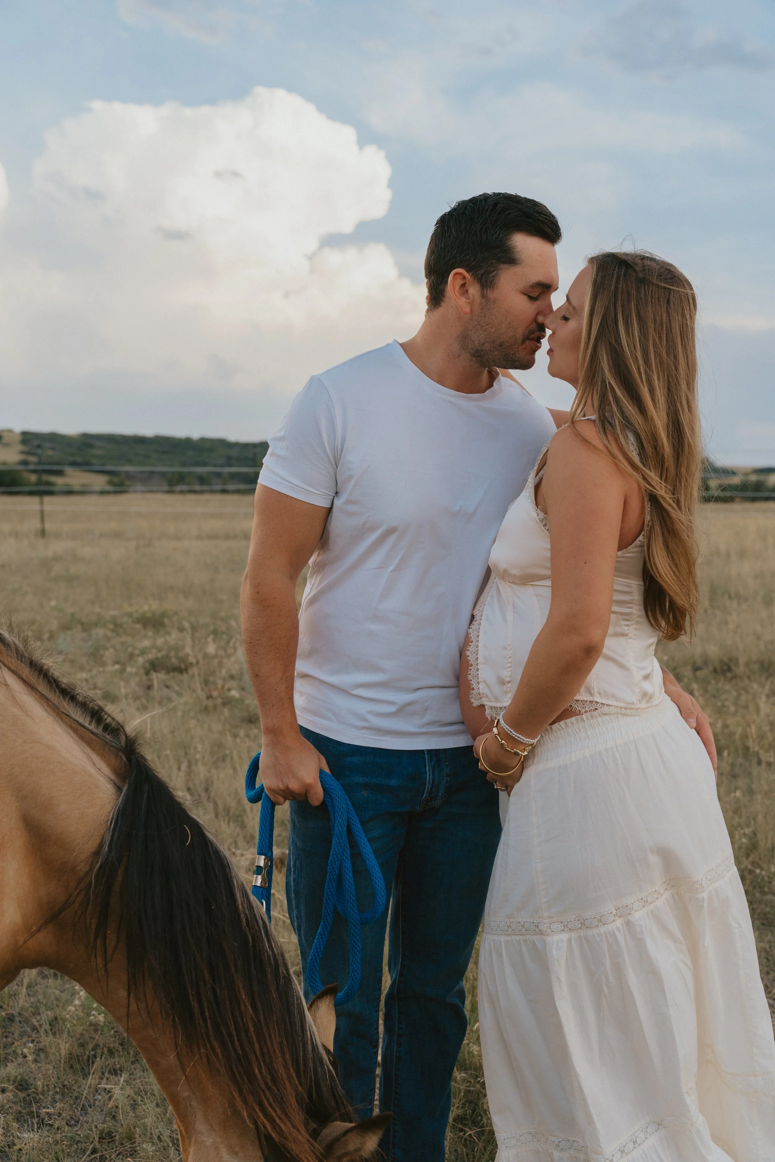 A couple kissing outdoors in a field, with a horse nearby, under a partly cloudy sky.