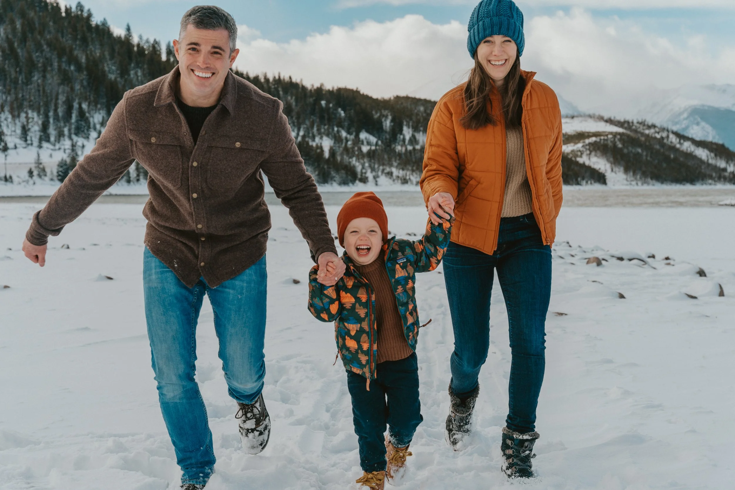 A family of three, two adults and a child, smiling and holding hands while walking in the snow near a lake with mountains in the background.