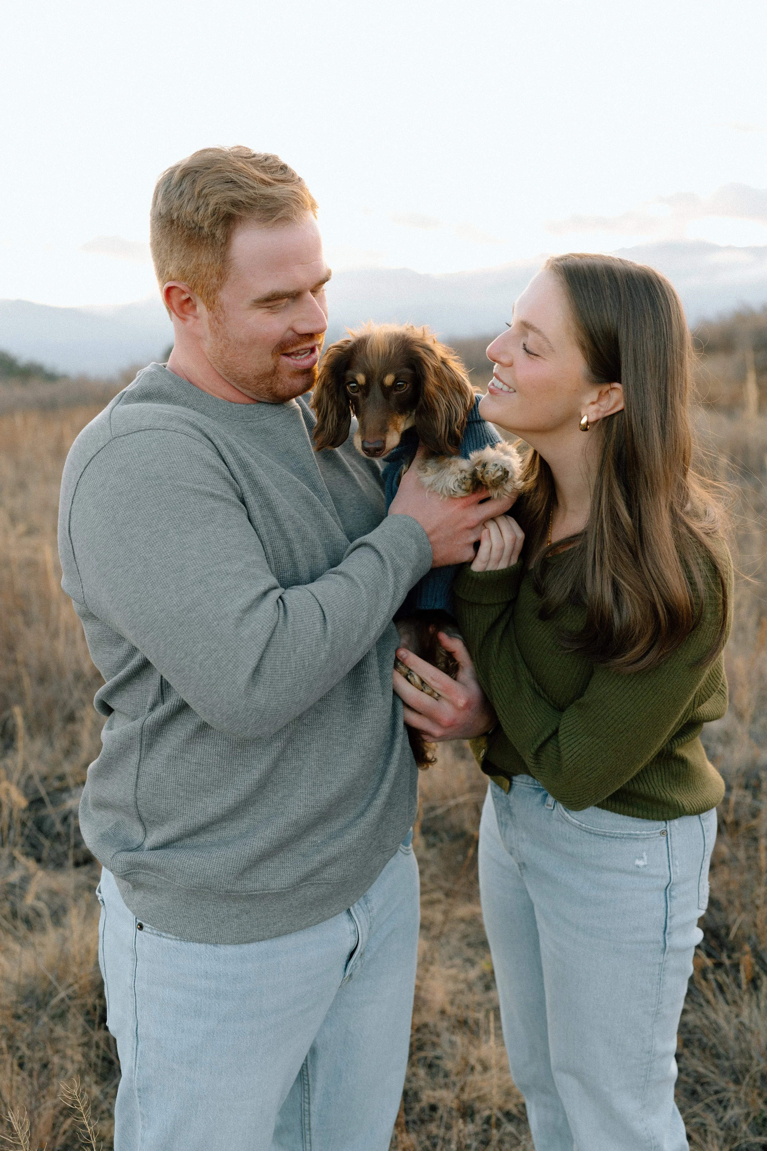 A couple holding a dachshund dog outdoors in a field during sunset, with mountains in the background.