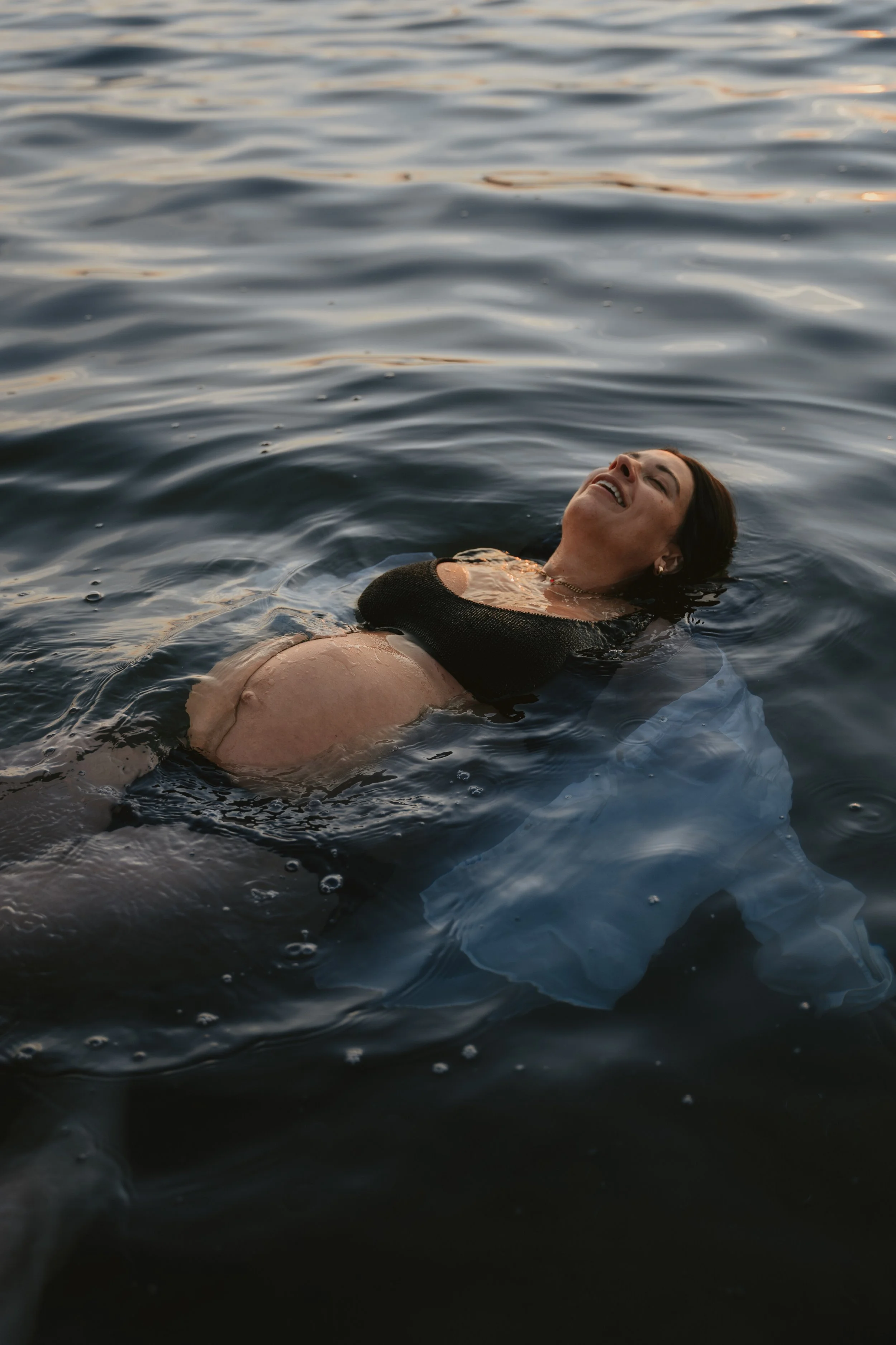 A woman floating in water, smiling with her head tilted back, wearing a black top and light-colored bottom, near sunset.