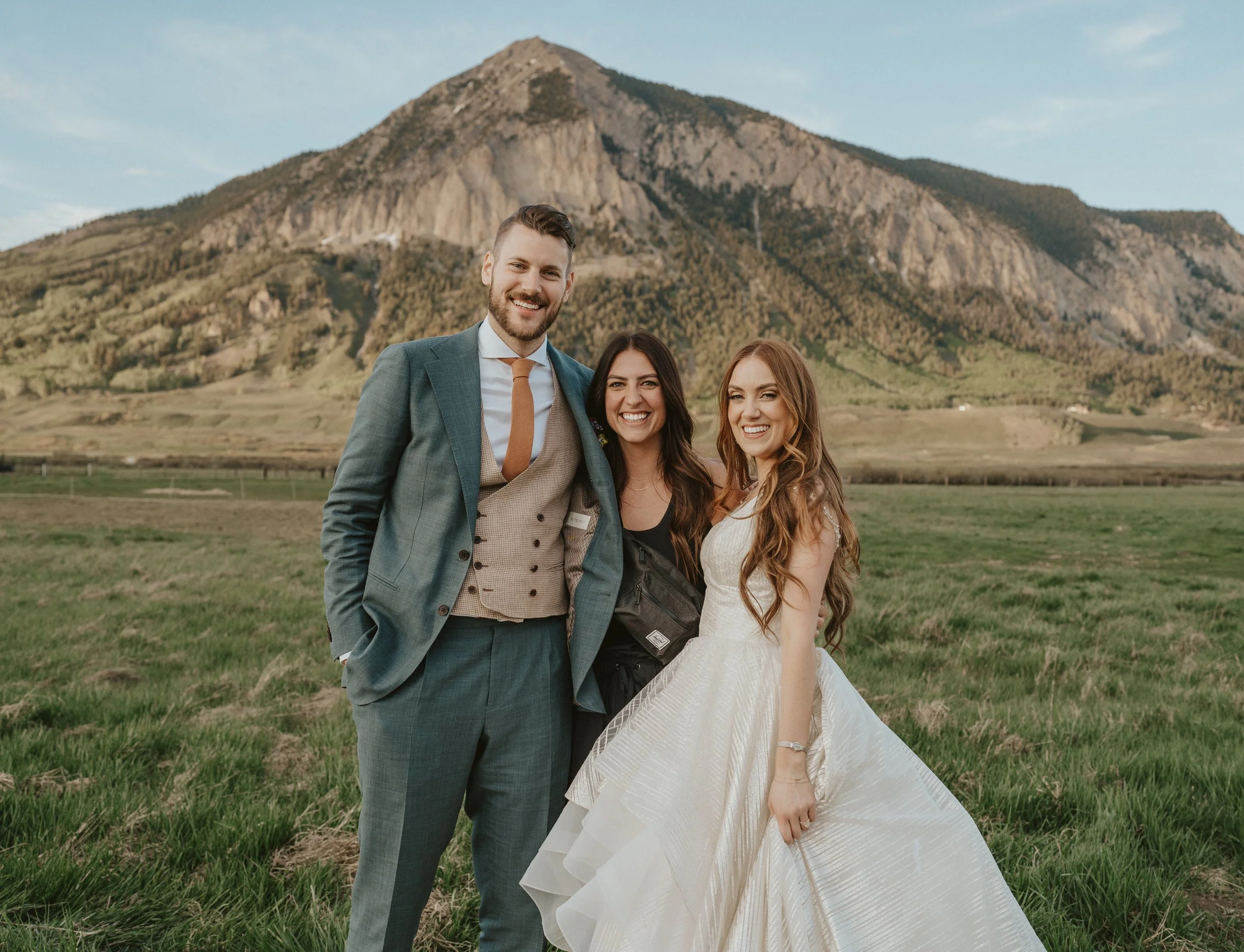 Three people smiling outdoors in front of a mountain, one man in a suit and two women, one in a wedding dress.