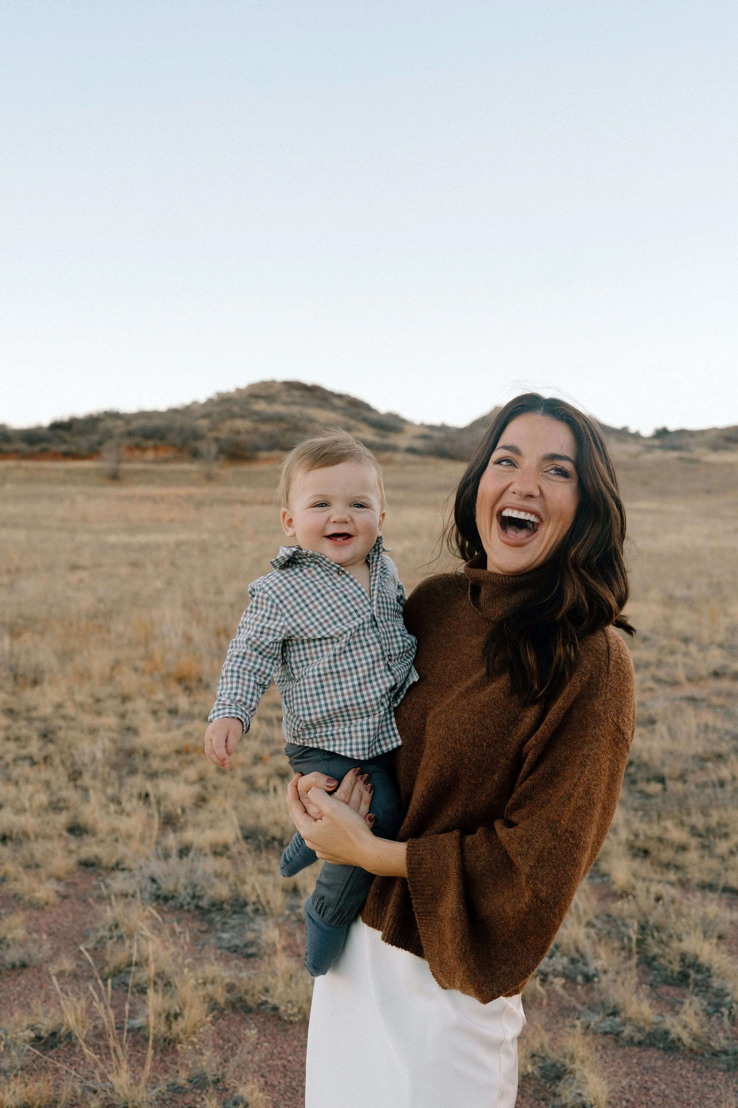A woman holding a smiling baby in an open field with hills in the background.