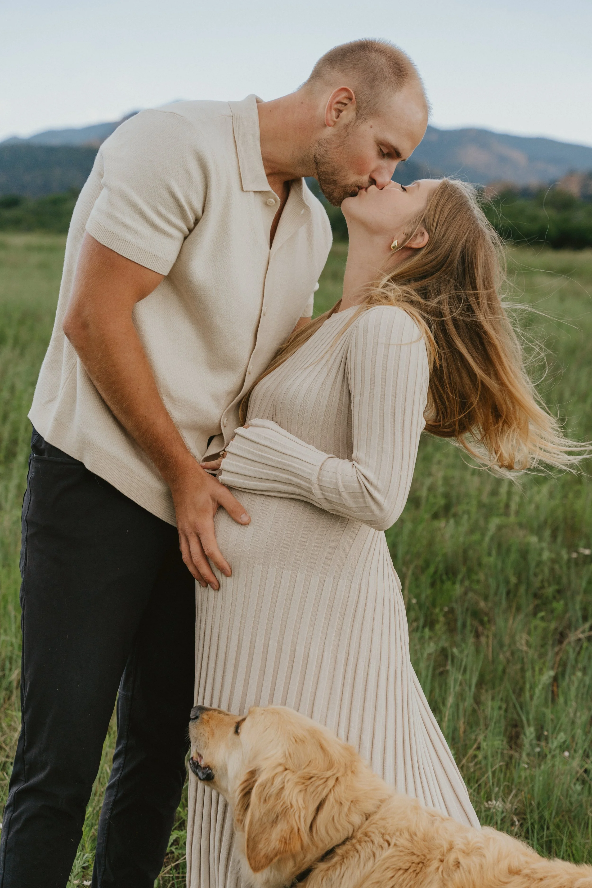 A couple kissing outdoors with a golden retriever puppy in a grassy field.