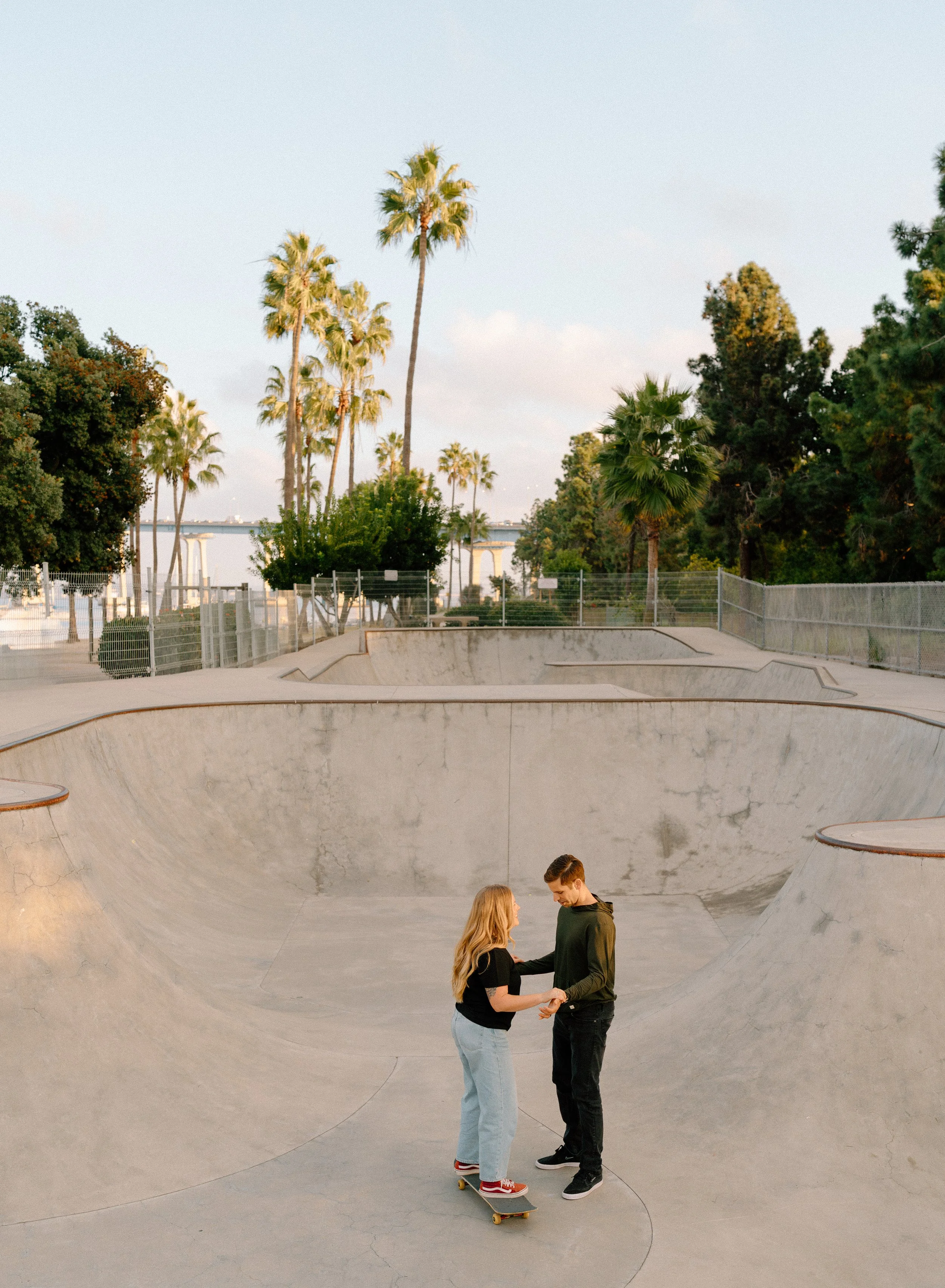 A young couple is skating and dancing together in an outdoor concrete skatepark with palm trees and fences in the background, under a clear sky.